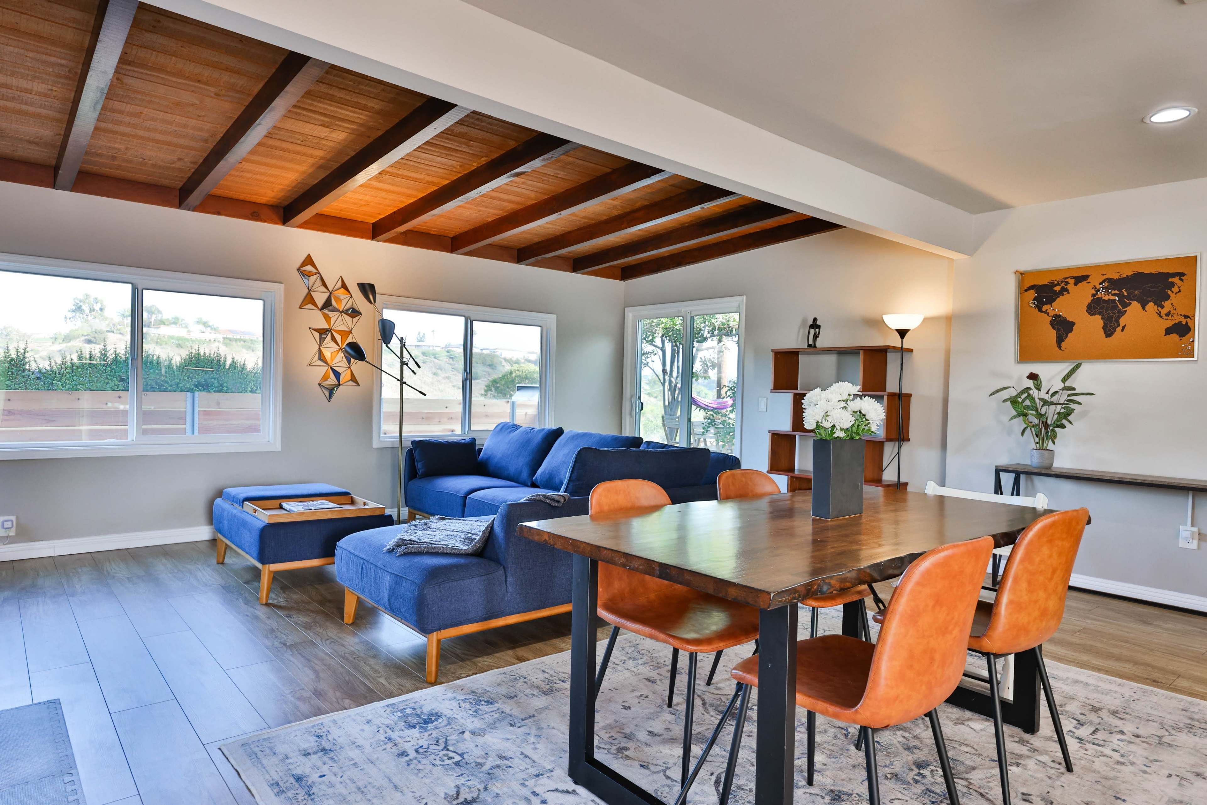 A modern living and dining area featuring a blue sofa, a wooden dining table with orange chairs, and large windows that allow natural light to fill the space.