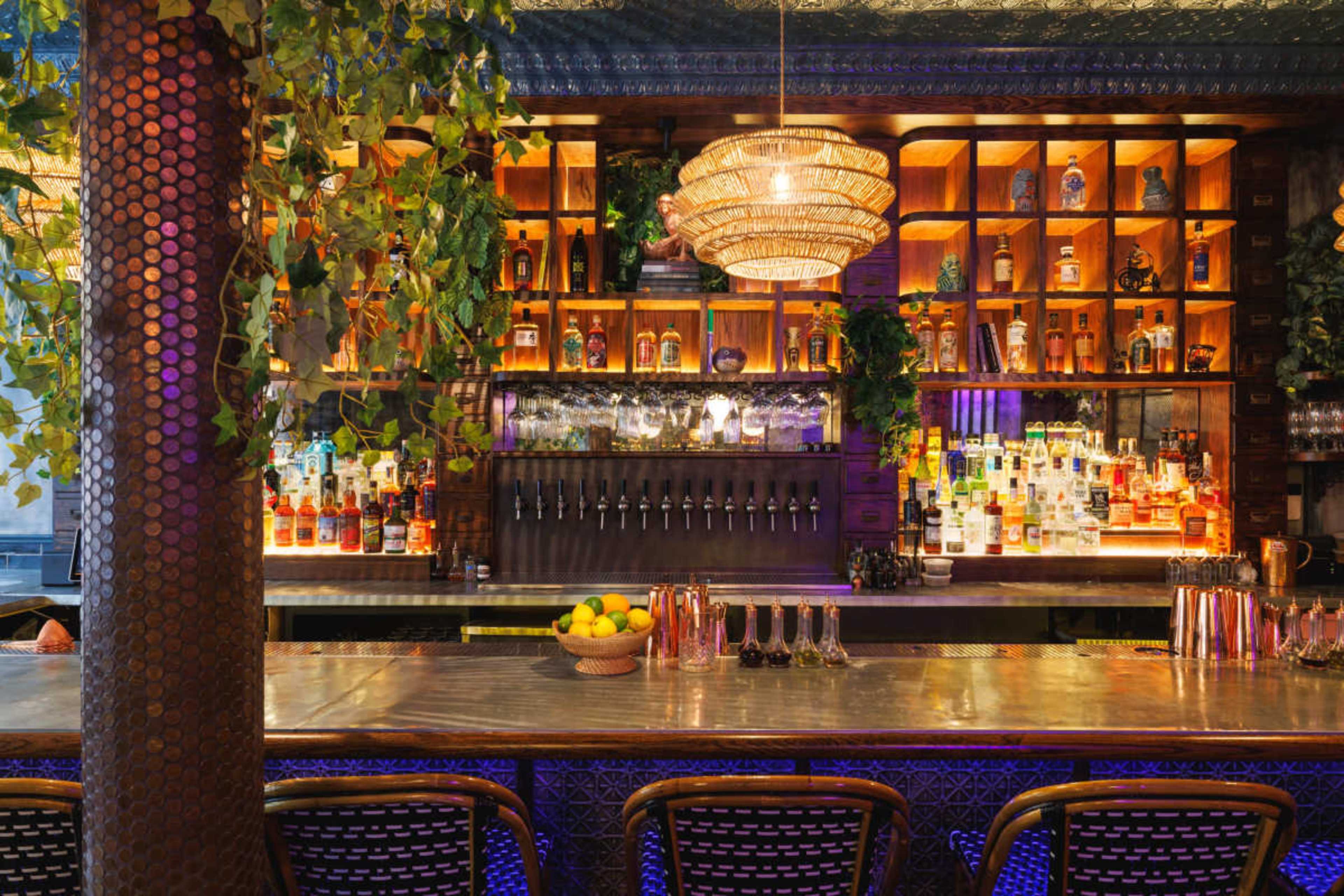 The image shows a well-lit bar featuring an array of colorful bottles on shelves behind a polished metal counter with a decorative chandelier overhead.
