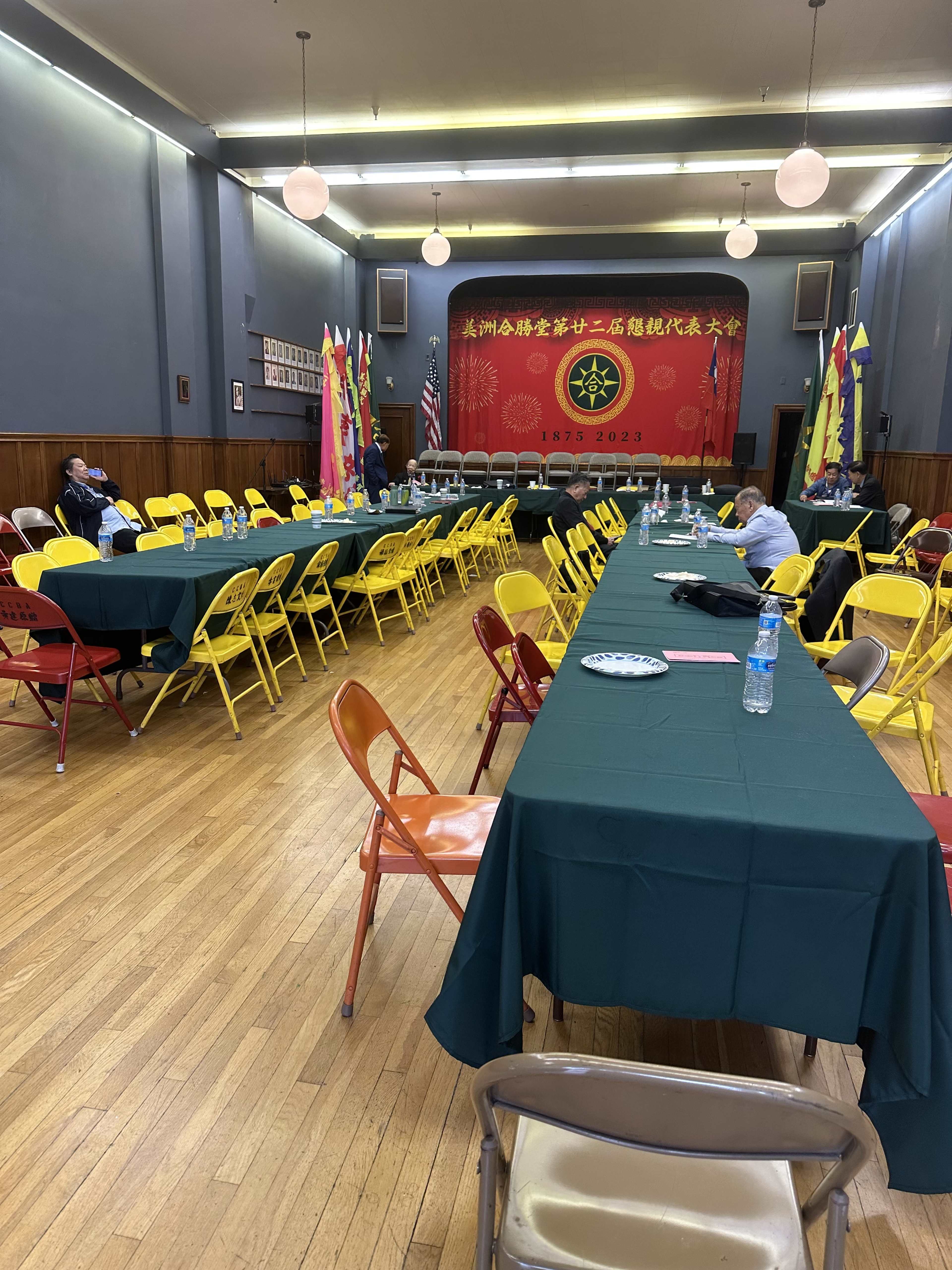 A large hall with yellow and green folding chairs arranged around several long tables, featuring a red backdrop with decorative banners and flags.