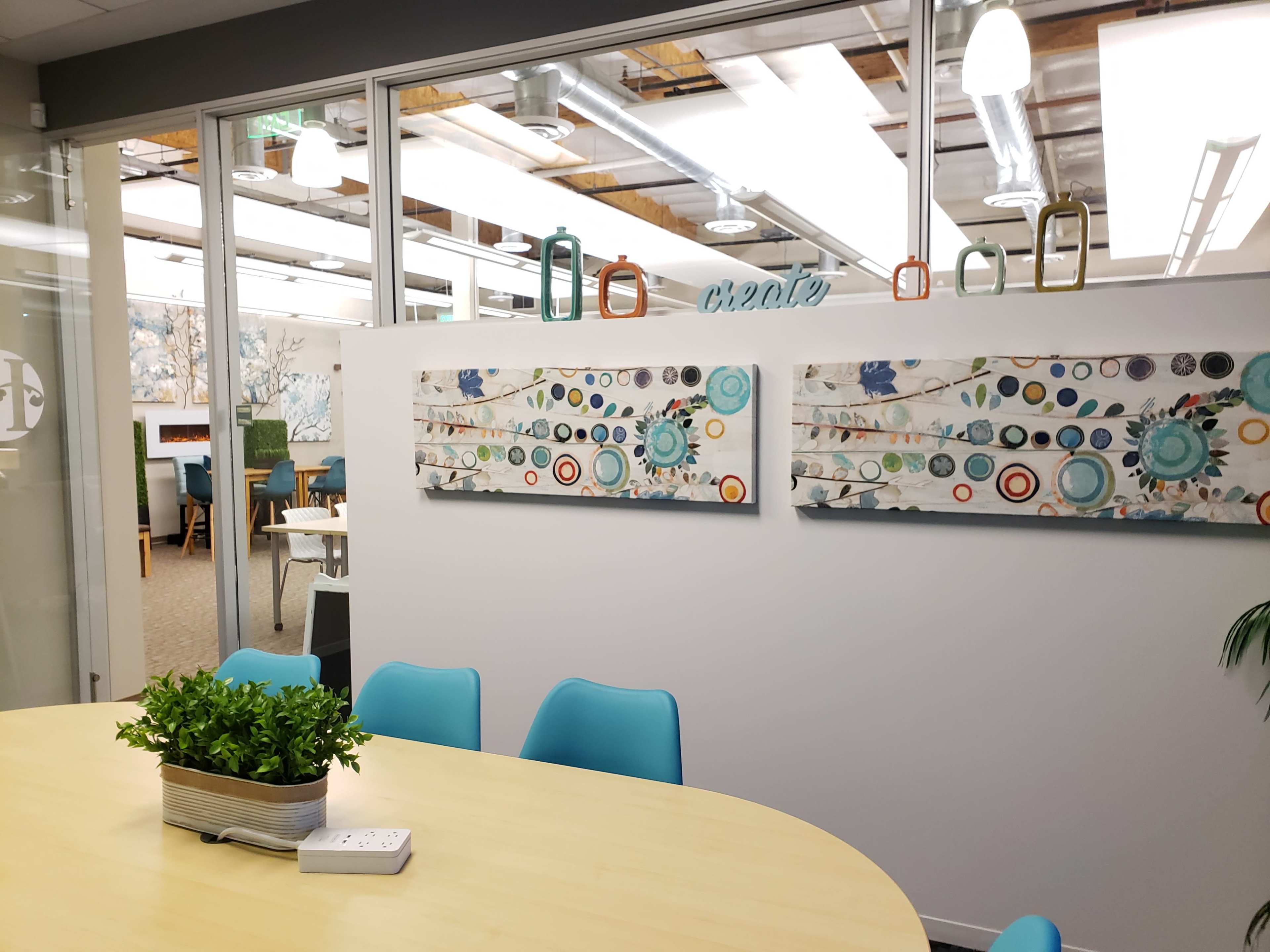 The image shows a modern office space with two decorative wall hangings, a round table with blue chairs, and greenery in a planter.