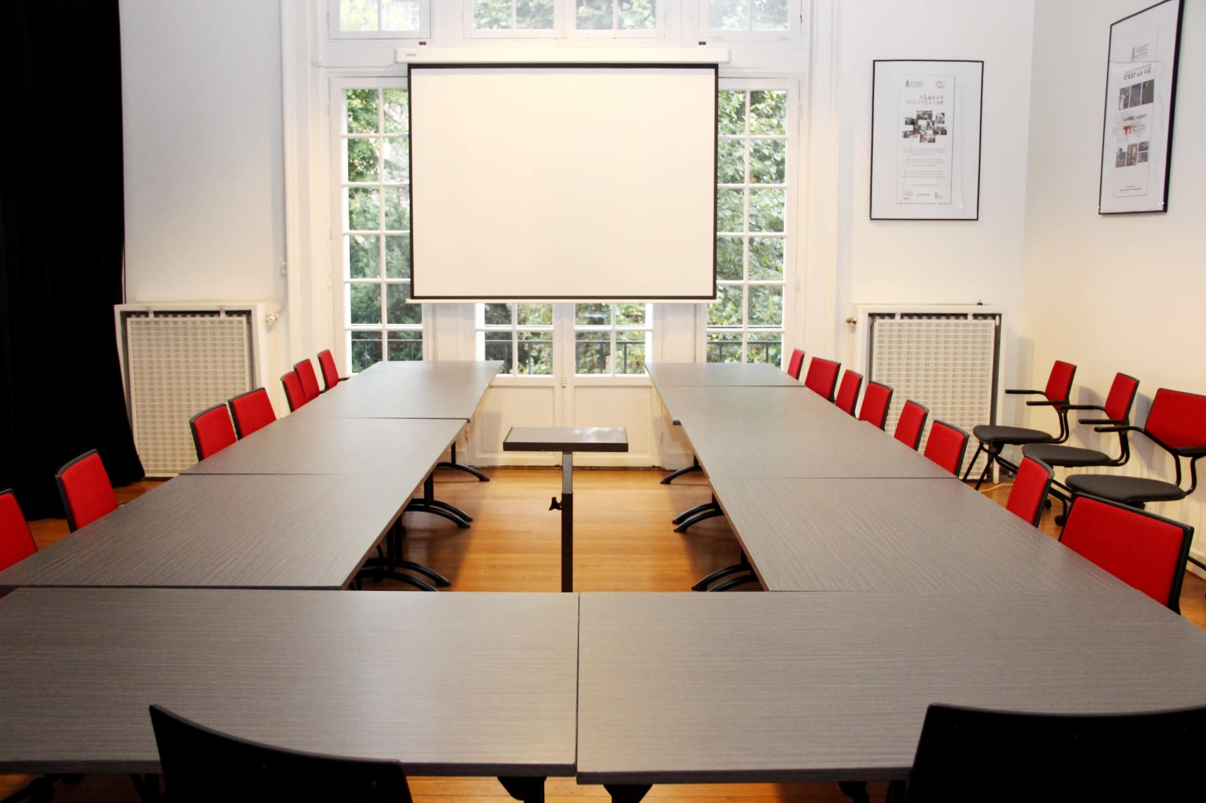 A large meeting room features two sets of rectangular tables arranged in a U-shape, surrounded by red chairs, with a blank projection screen at one end and large windows providing natural light.