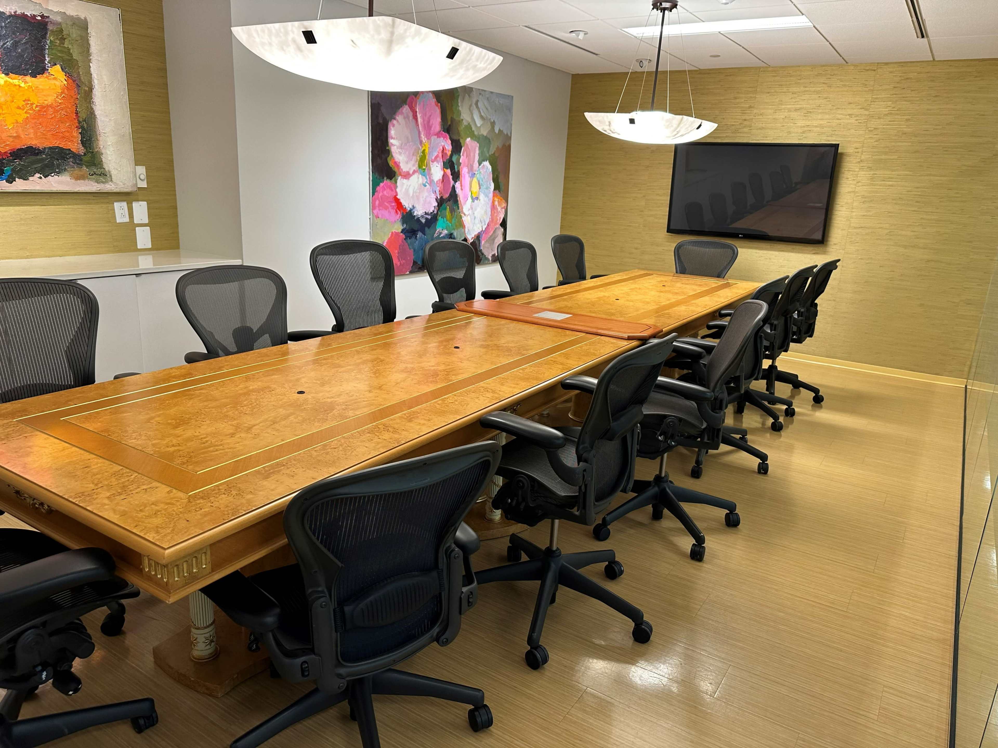 A modern conference room featuring a large wooden table surrounded by black office chairs, with artwork on the walls and a wall-mounted screen.