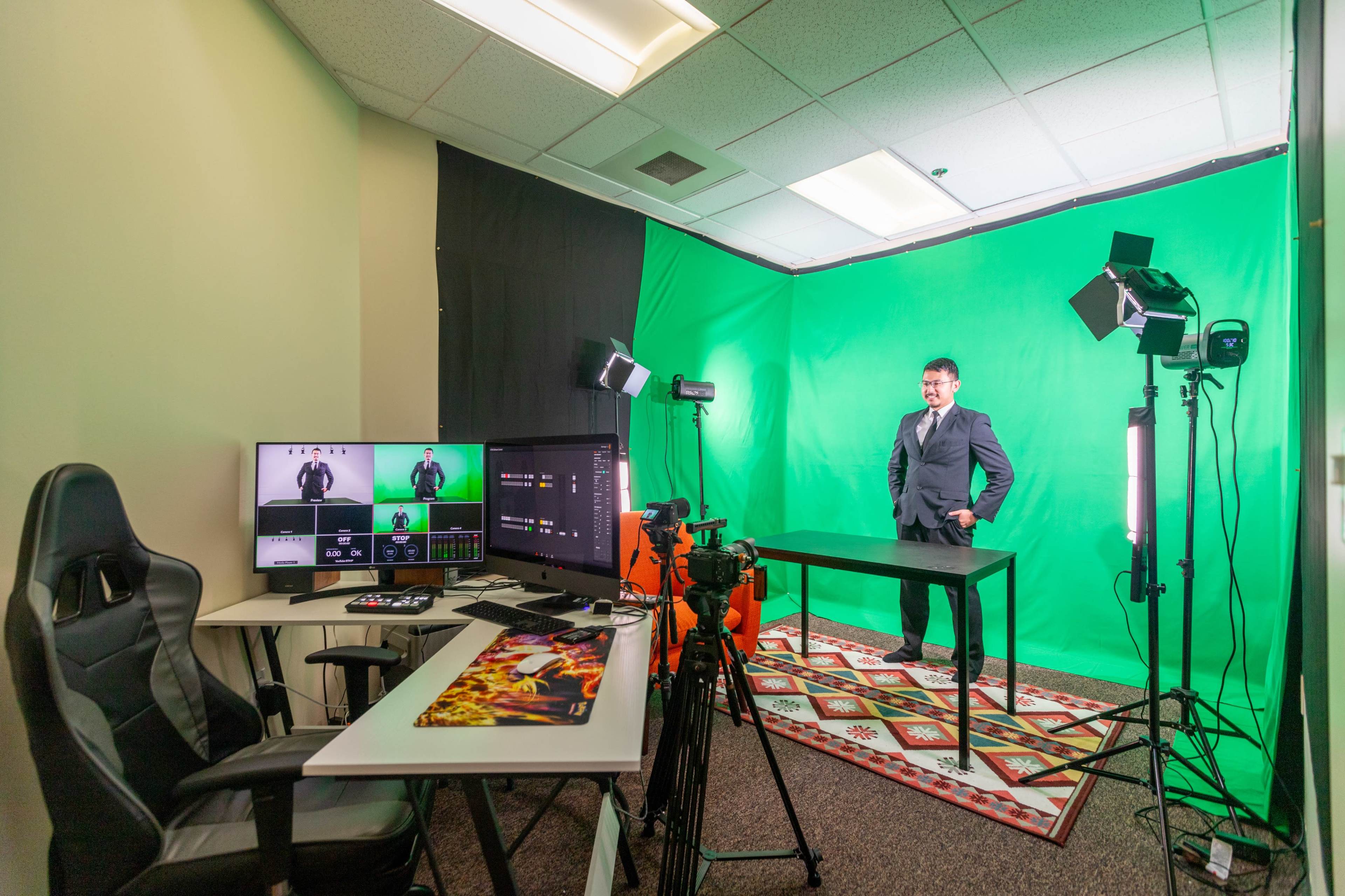A green screen setup for video production features a man in formal attire, a desk, video equipment, and multiple monitors displaying live feeds.