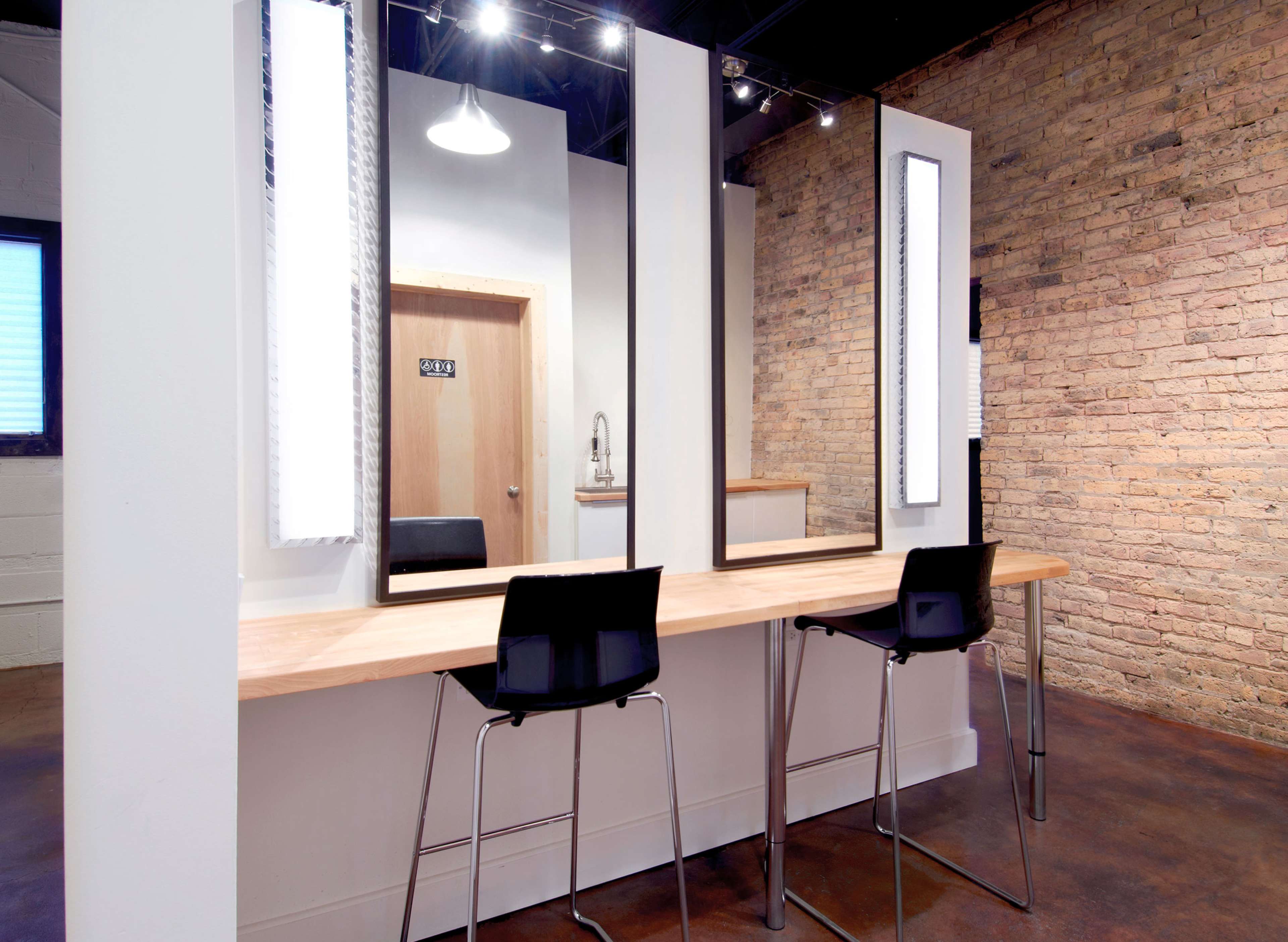 A well-lit salon area with two black chairs and three vertical mirrors mounted on a light-colored wall, accentuated by exposed brick.