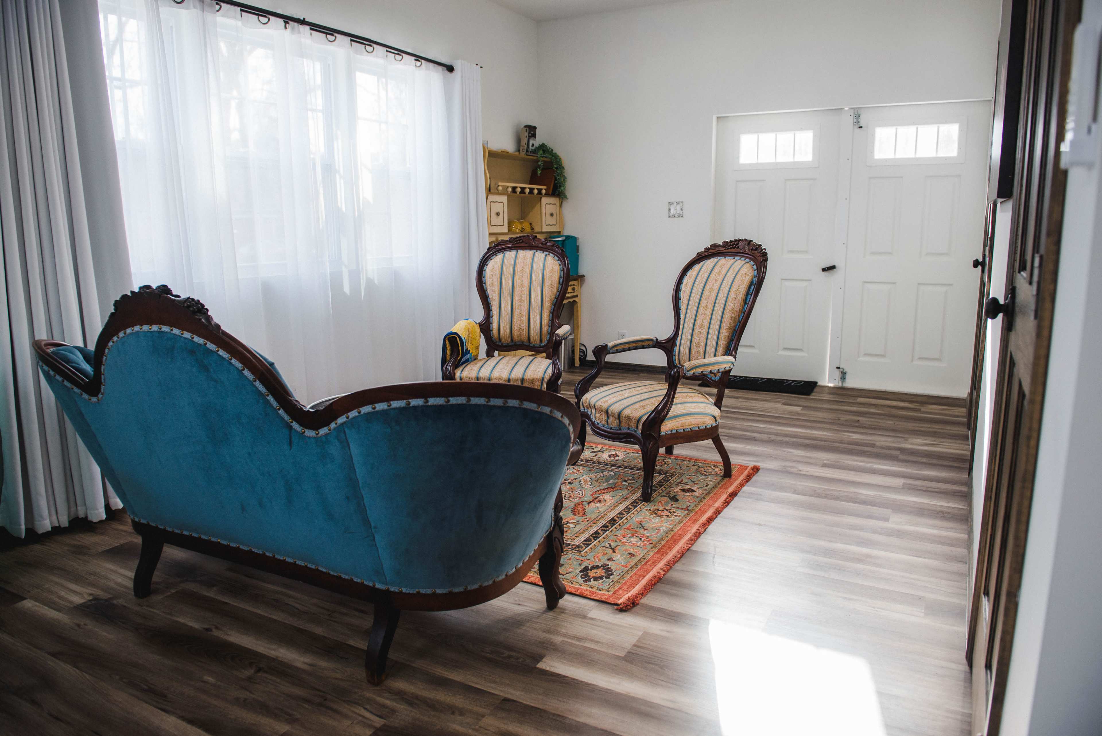 The scene shows a living room with two upholstered chairs and a couch, all arranged atop a patterned rug, leading to a pair of double doors.