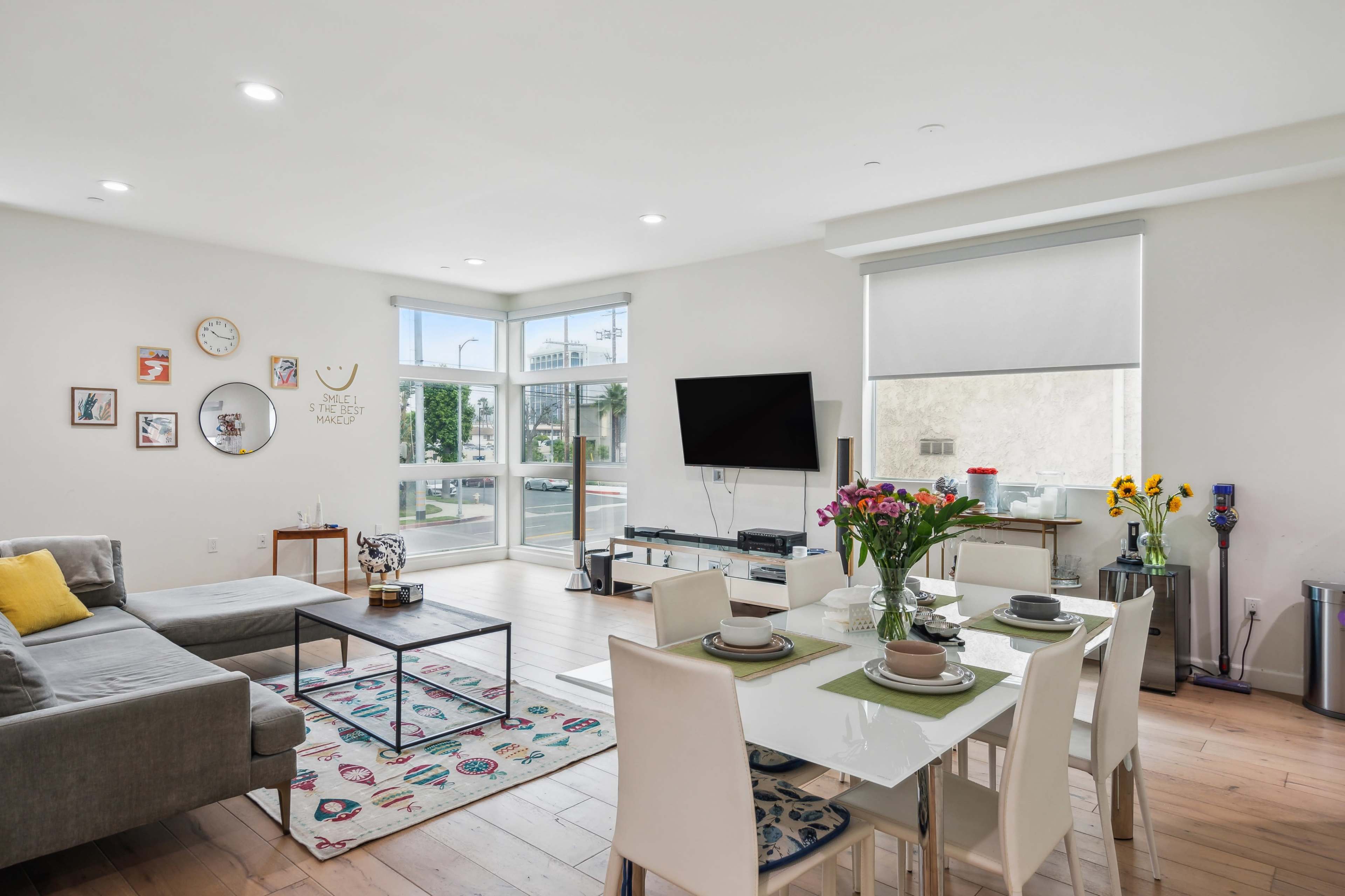 A modern living space features a gray sectional sofa, a dining table set for four, and large windows allowing natural light to fill the room.