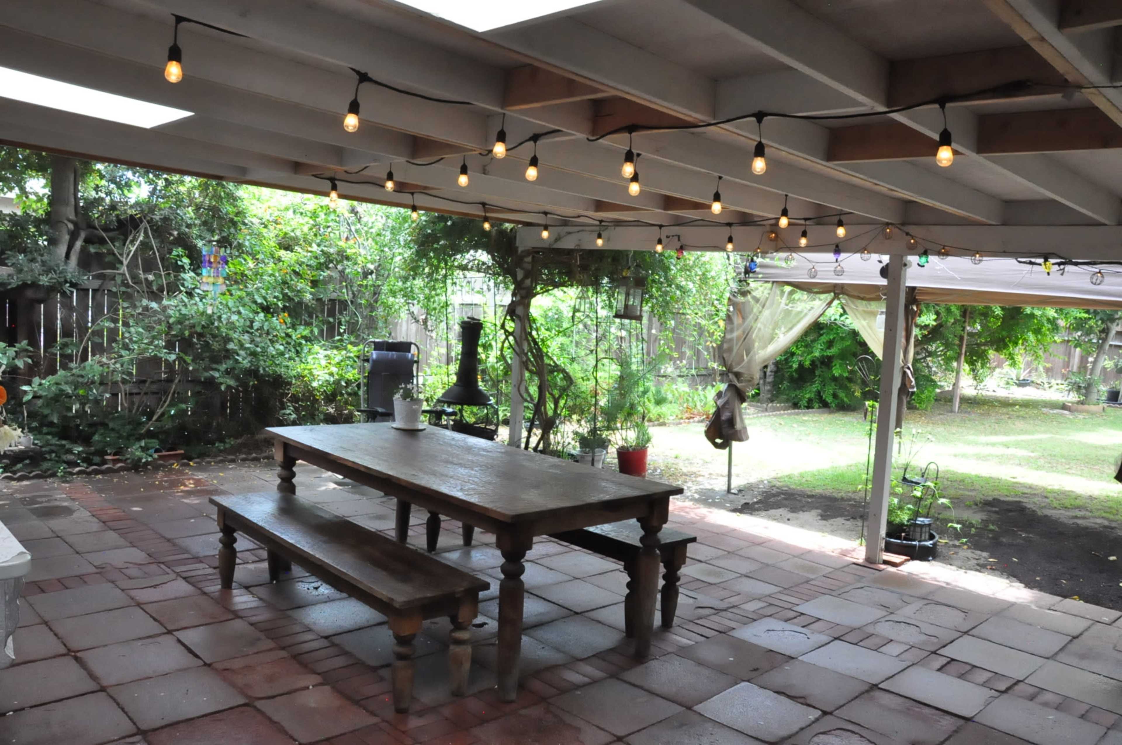 A covered outdoor patio with a long wooden table, string lights overhead, and a garden area in the background.