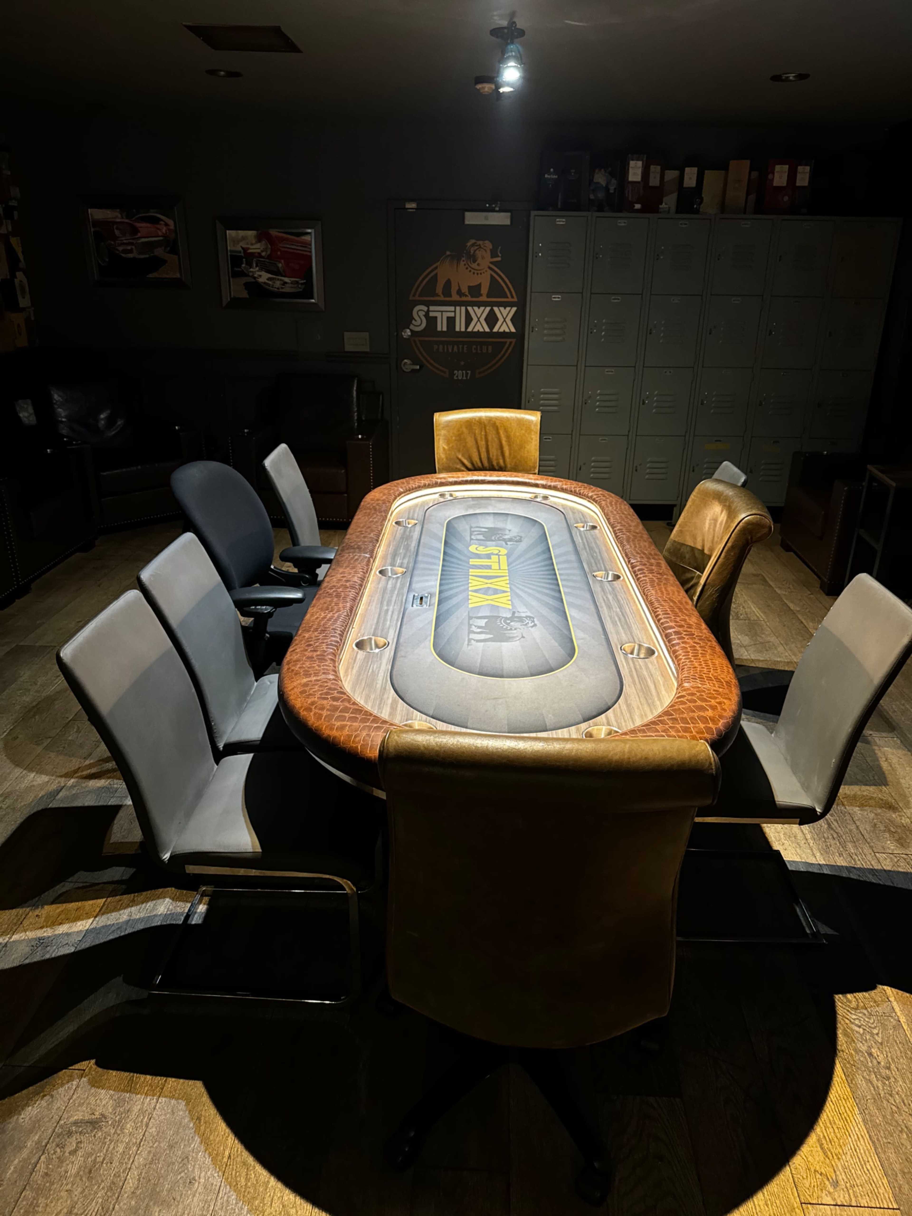 The image shows a poker table surrounded by chairs in a dimly lit room with lockers in the background.