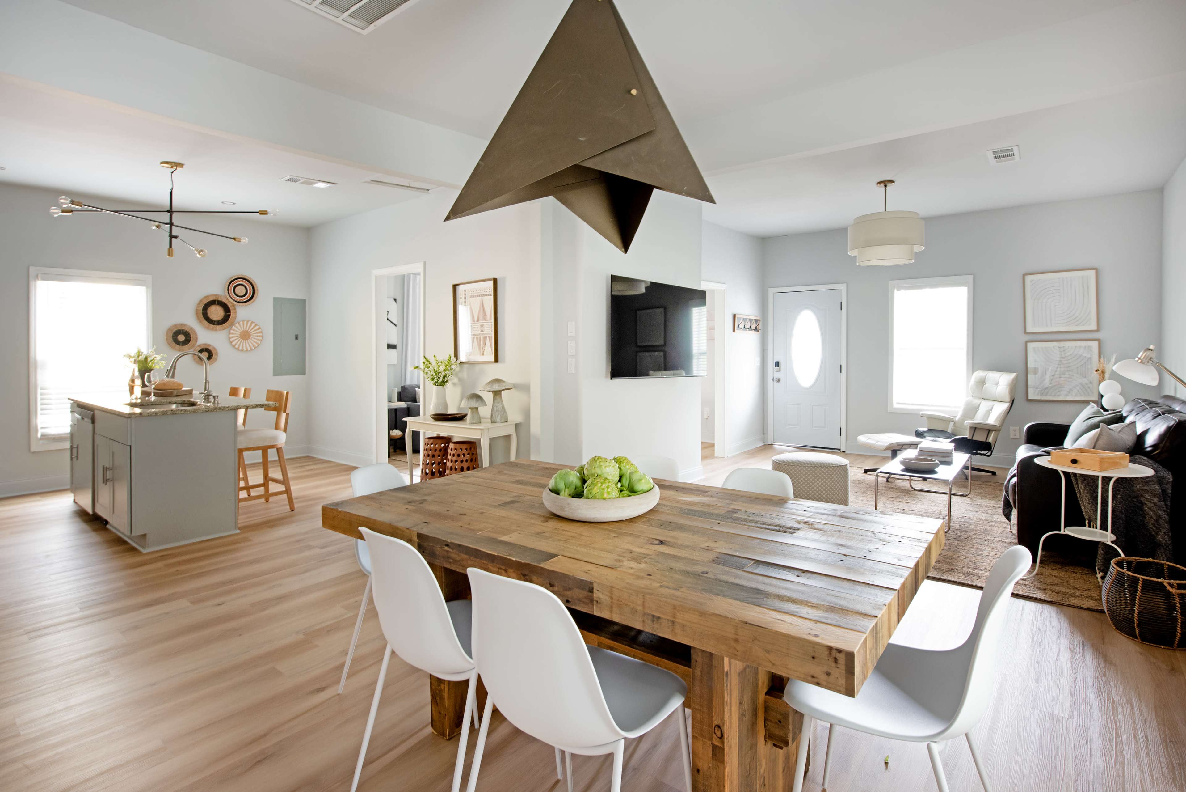 The image shows a modern open-concept living and dining area featuring a wooden dining table, white chairs, an island kitchen, and a cozy seating area with a television.
