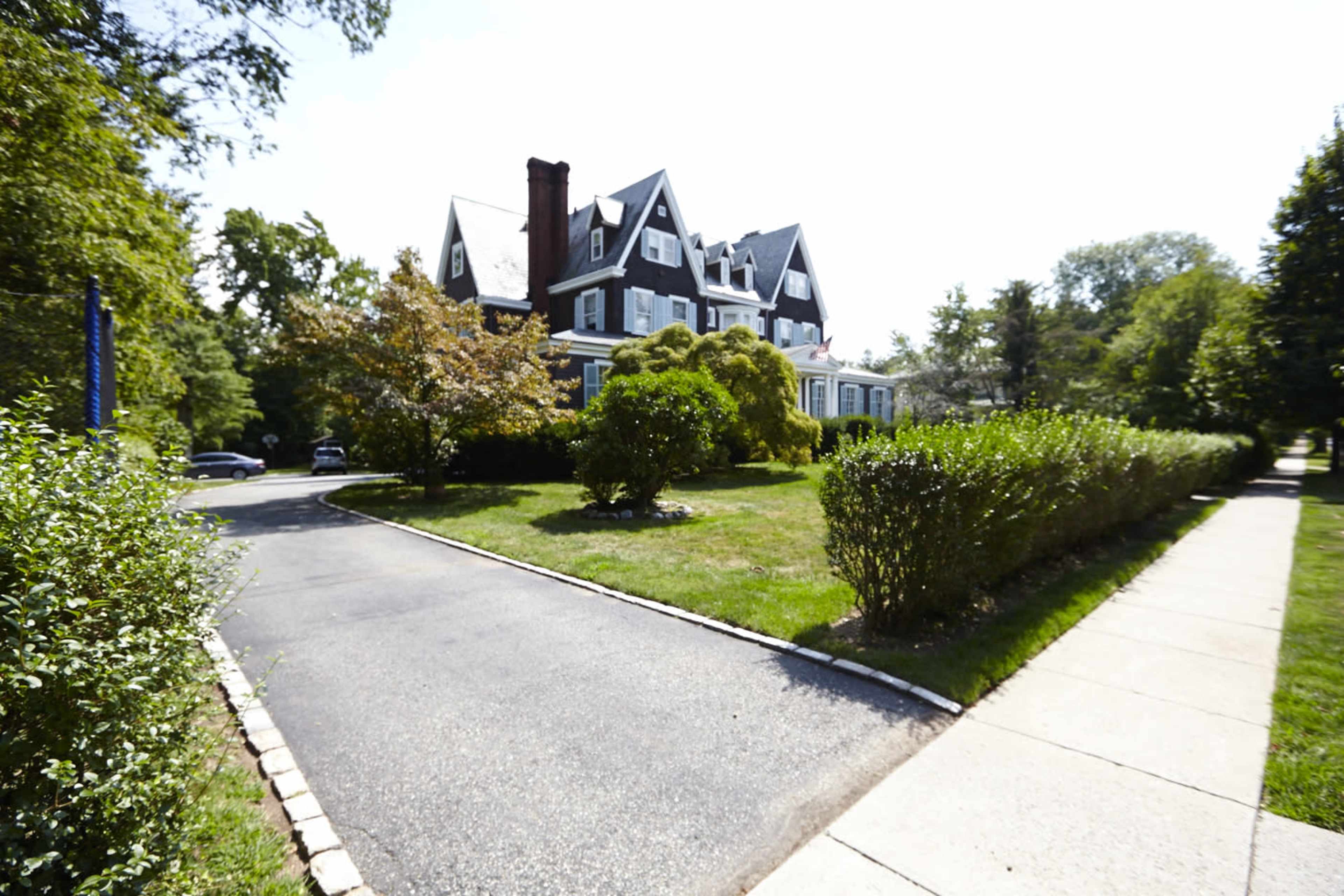 A large, multi-story house with a steeply pitched roof is set back from a tree-lined street, surrounded by manicured lawns and hedges.