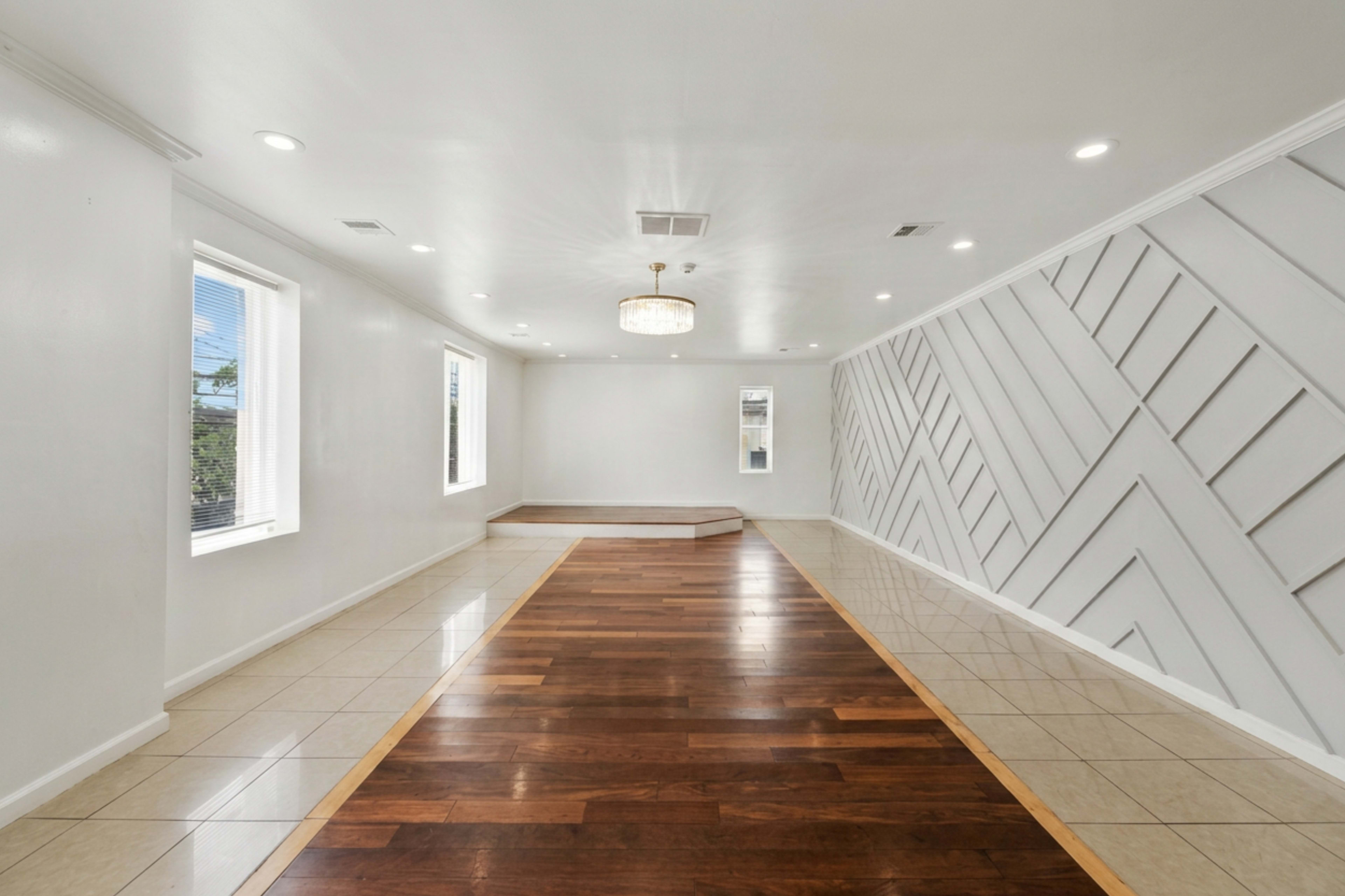 The image shows a spacious, open room with a mix of tiled and wooden flooring, decorated with textured wall panels and illuminated by natural light from windows.
