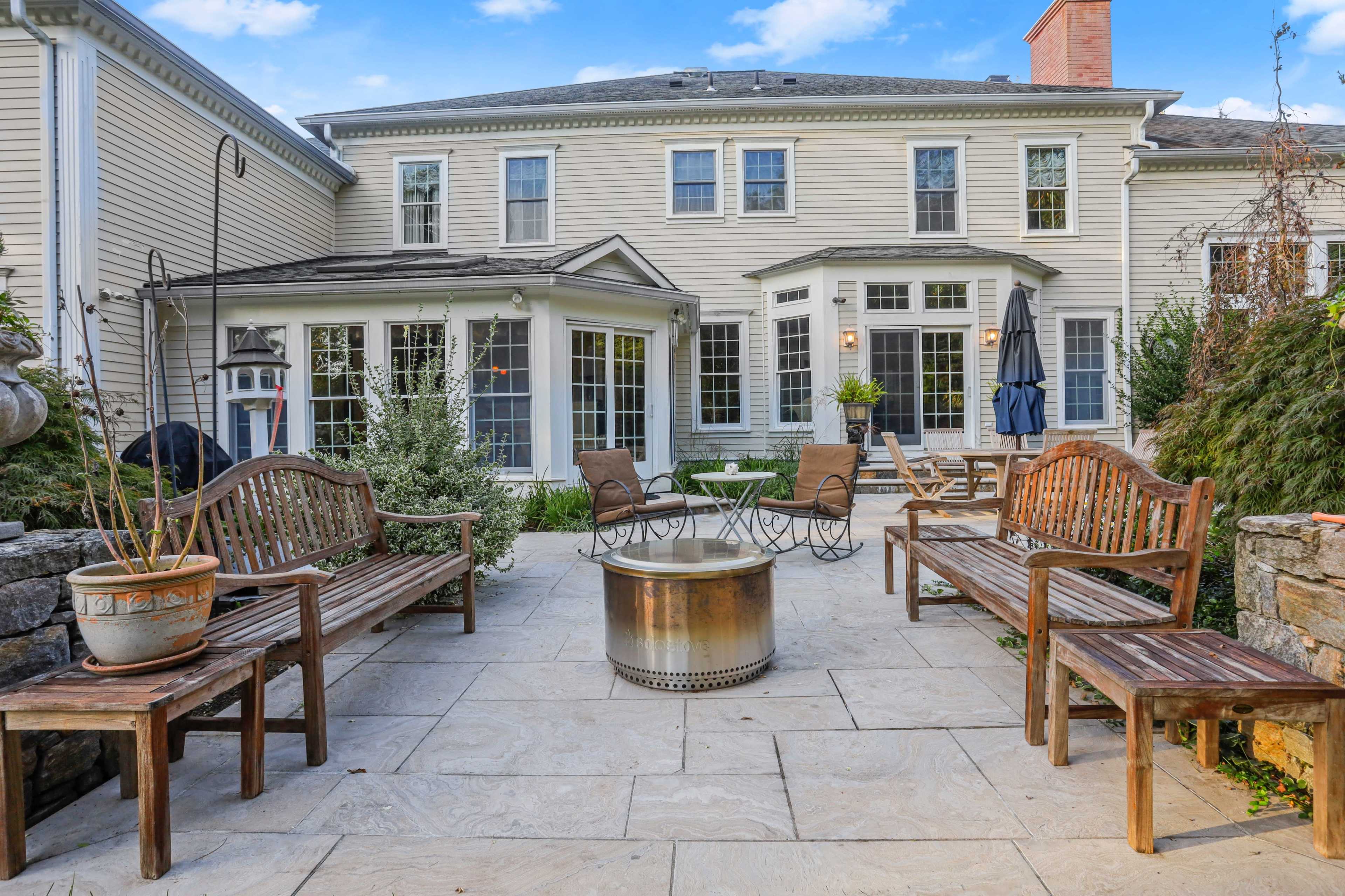 The image shows a stone patio area surrounded by wooden benches and a fire pit, with a house featuring multiple windows in the background.