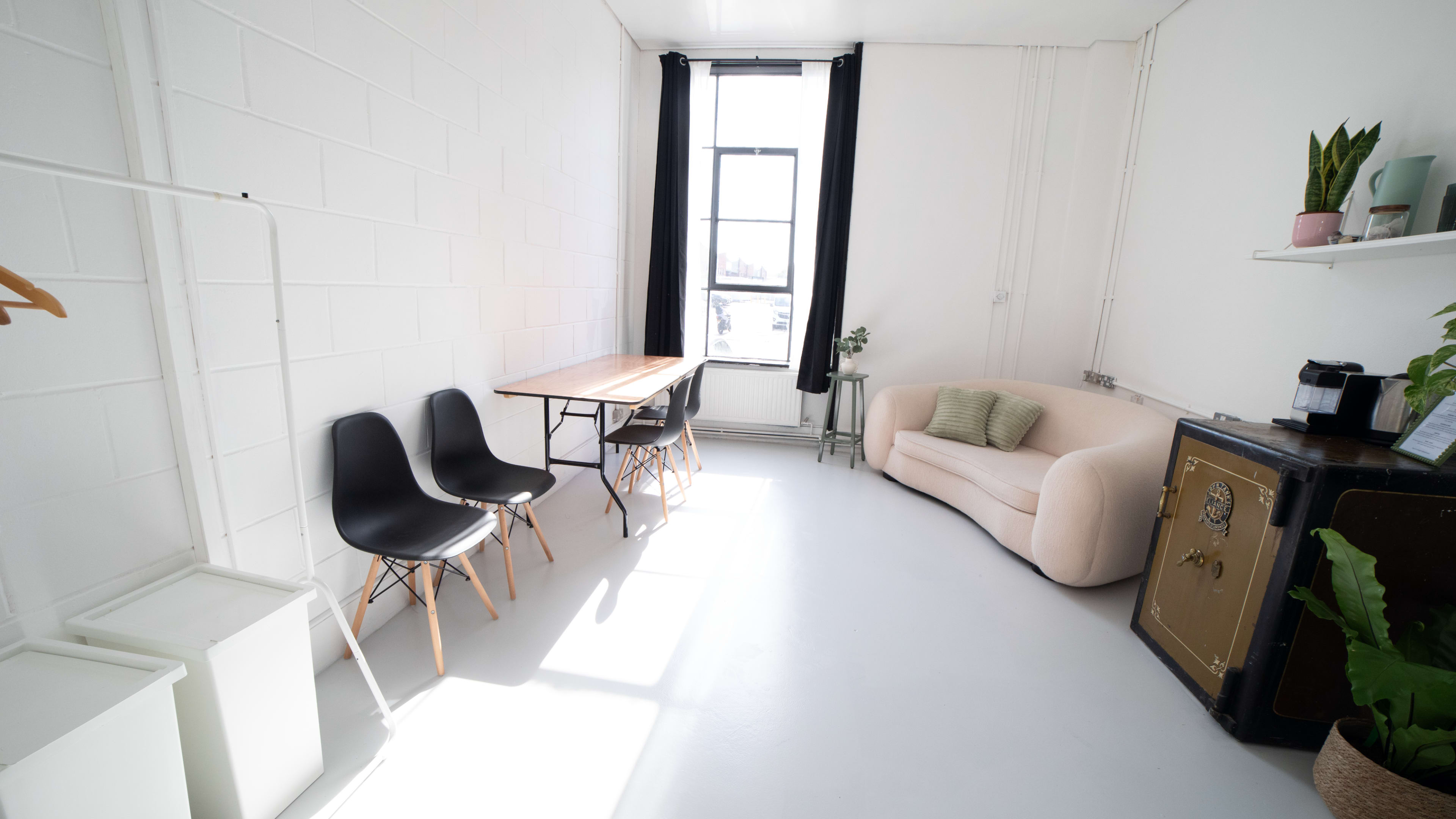 A bright, minimalistic room featuring a sofa, a wooden table with chairs, a plant, and a window.