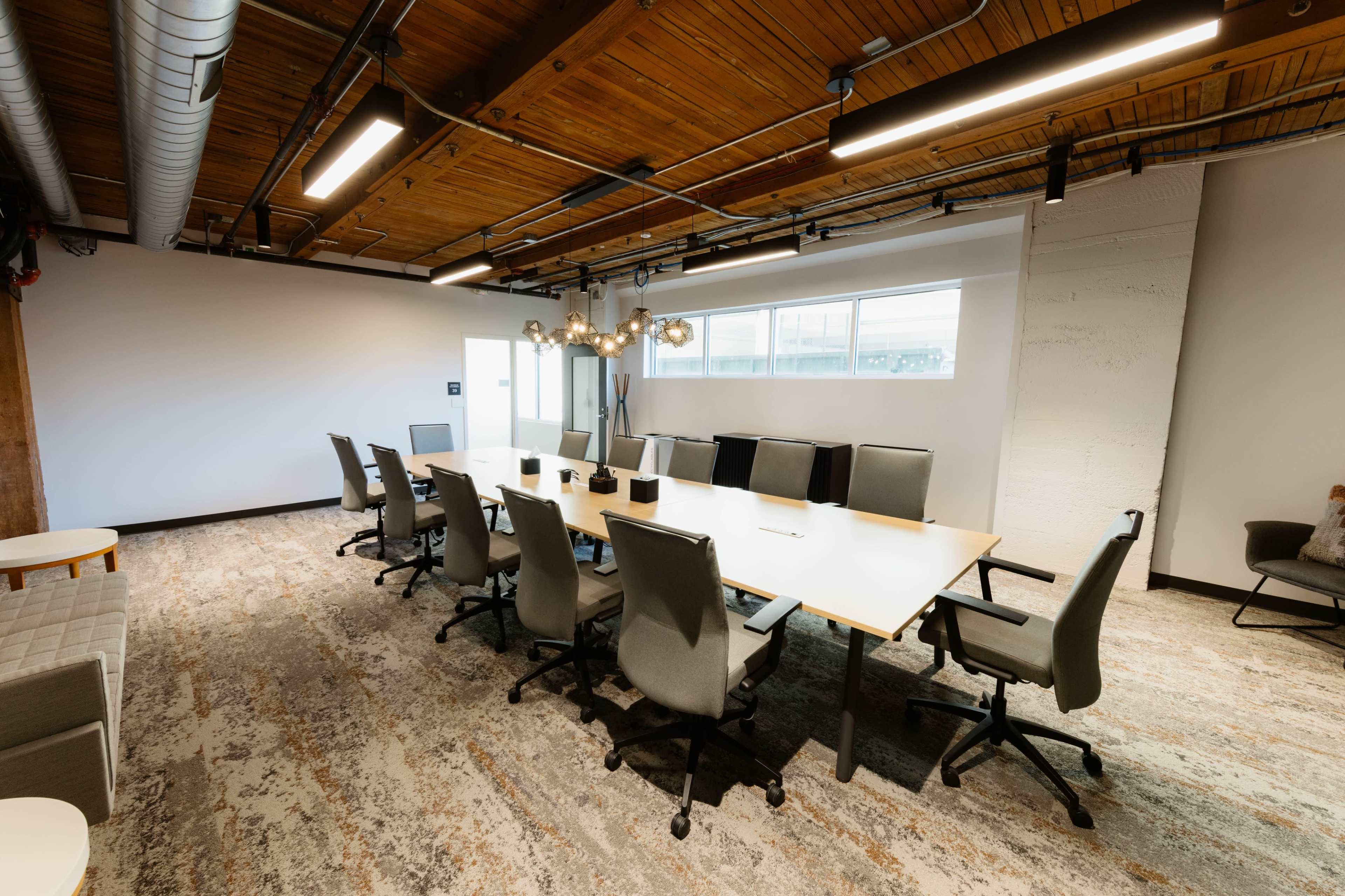 The image shows a modern conference room featuring a long white table surrounded by ergonomic chairs, with large windows providing natural light and wooden ceiling beams overhead.