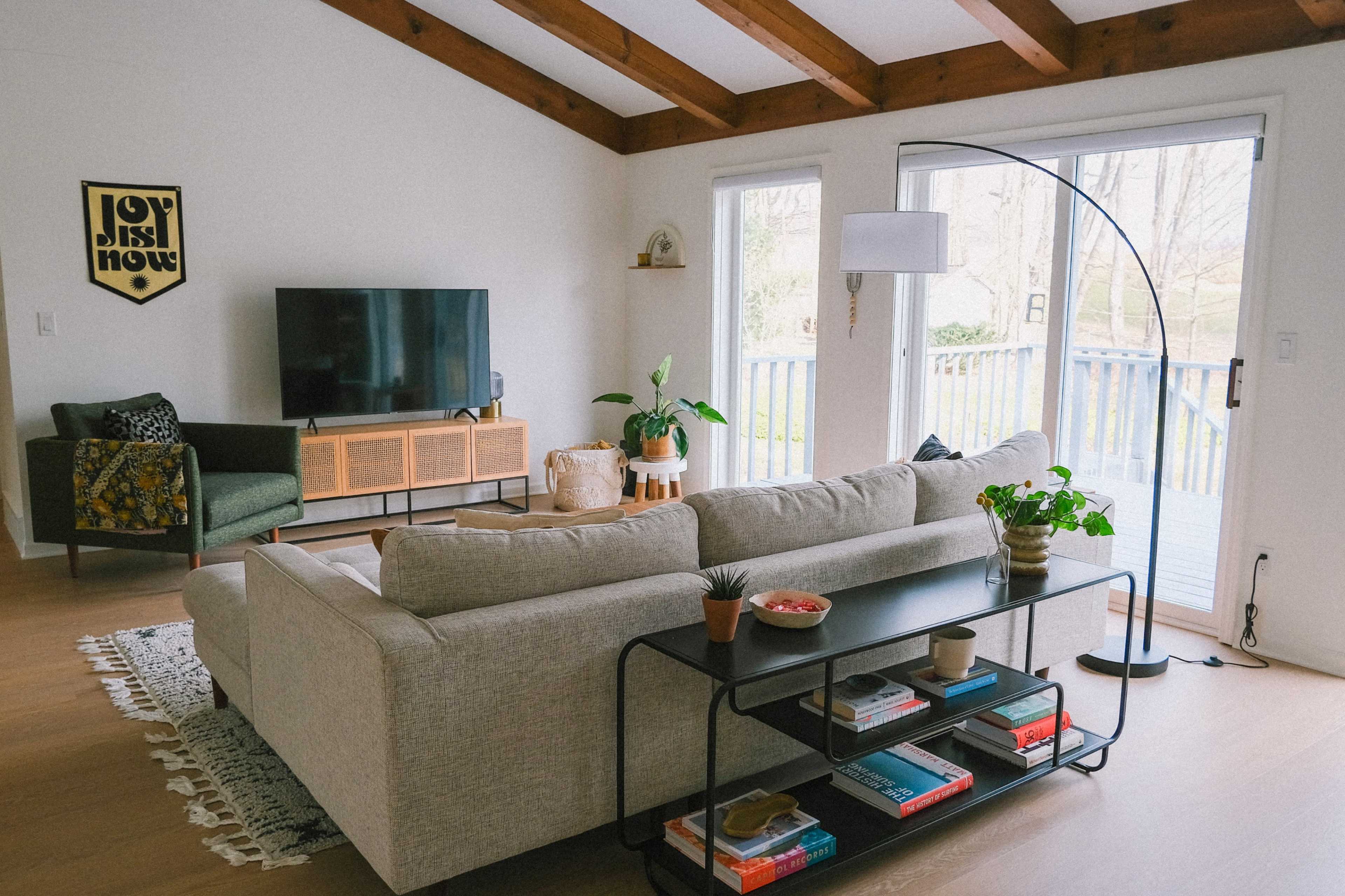 A modern living room features a light-filled space with a gray sectional sofa, a wooden TV stand, and large glass doors leading to an outdoor area.