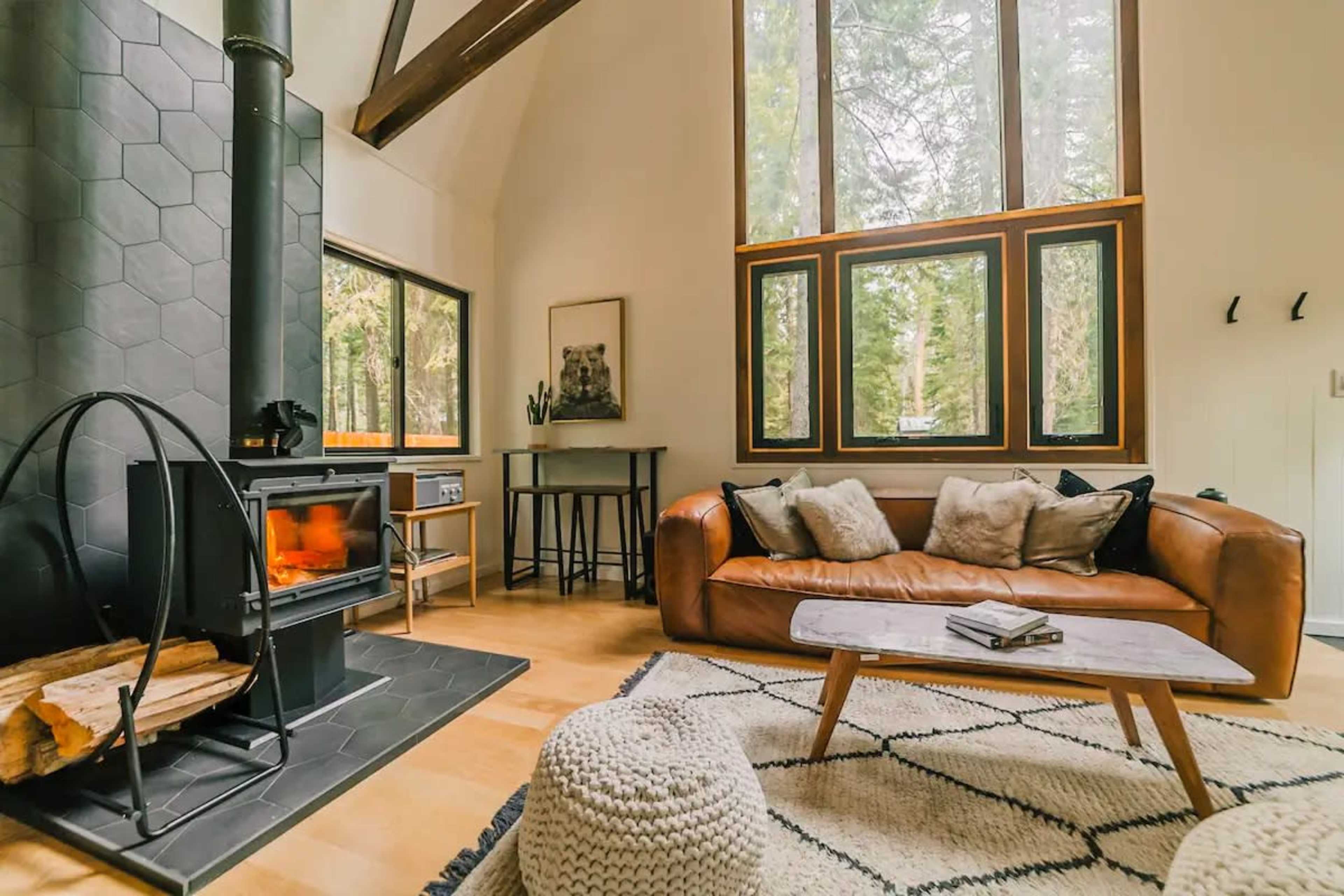 A cozy living room with a leather sofa, a stone fireplace, large windows allowing natural light, and a wooden coffee table on a patterned rug.