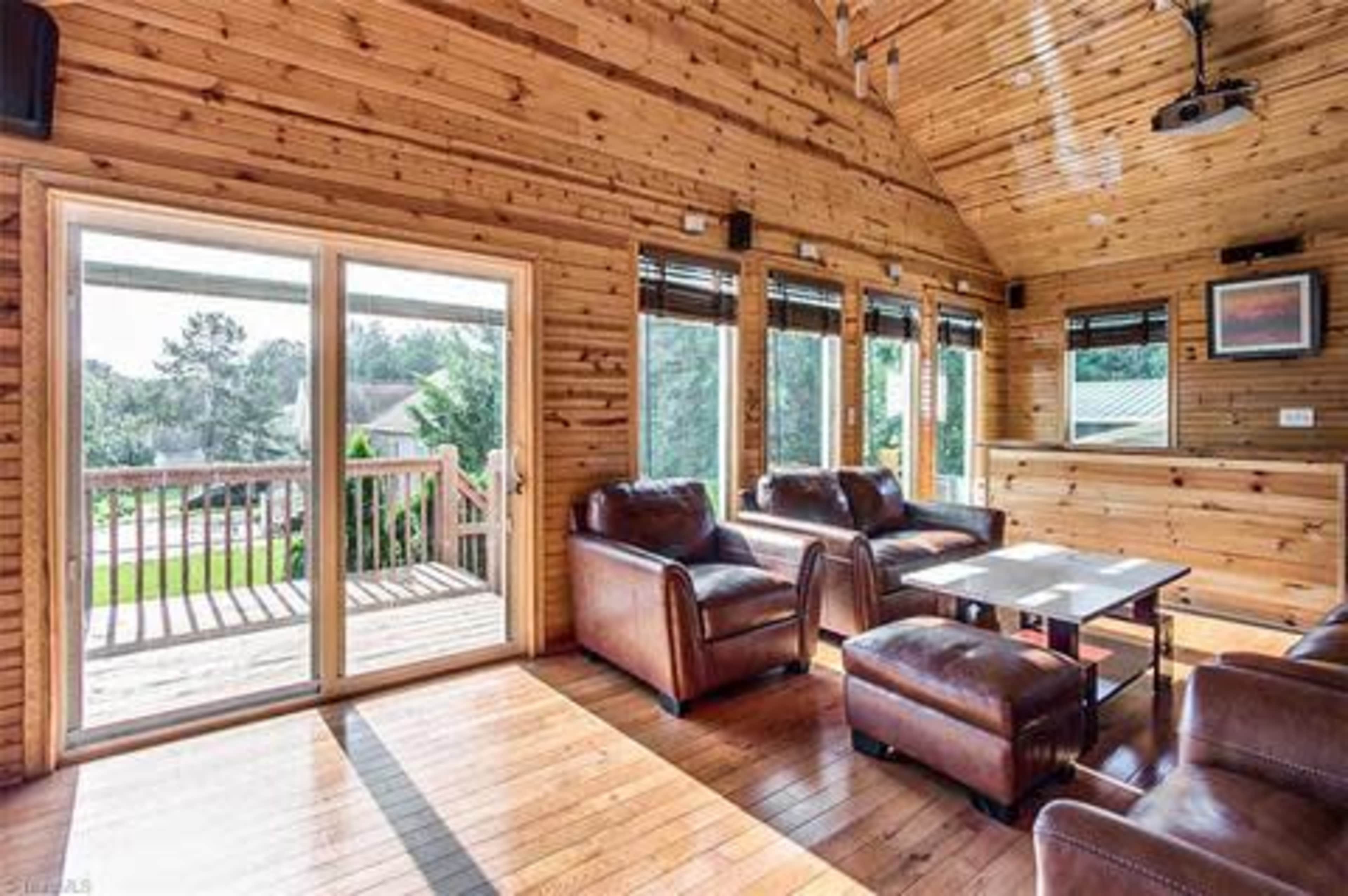 The interior of a wooden-paneled living room features leather couches, a coffee table, and large windows with a view of the outdoors.