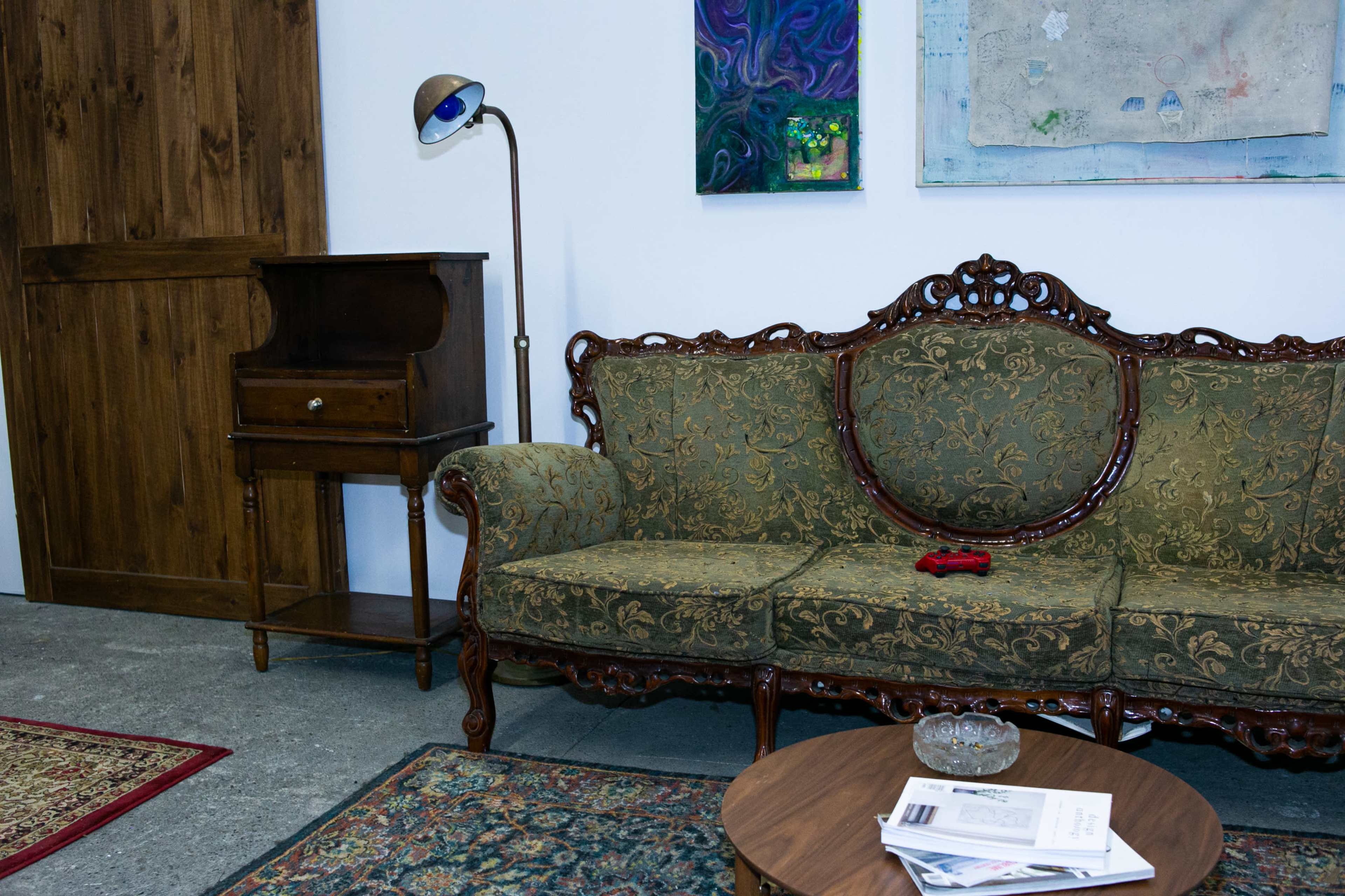 A vintage sofa with ornate carvings sits beside a wooden cabinet and a round table on a patterned rug, with a game controller resting on the seat.