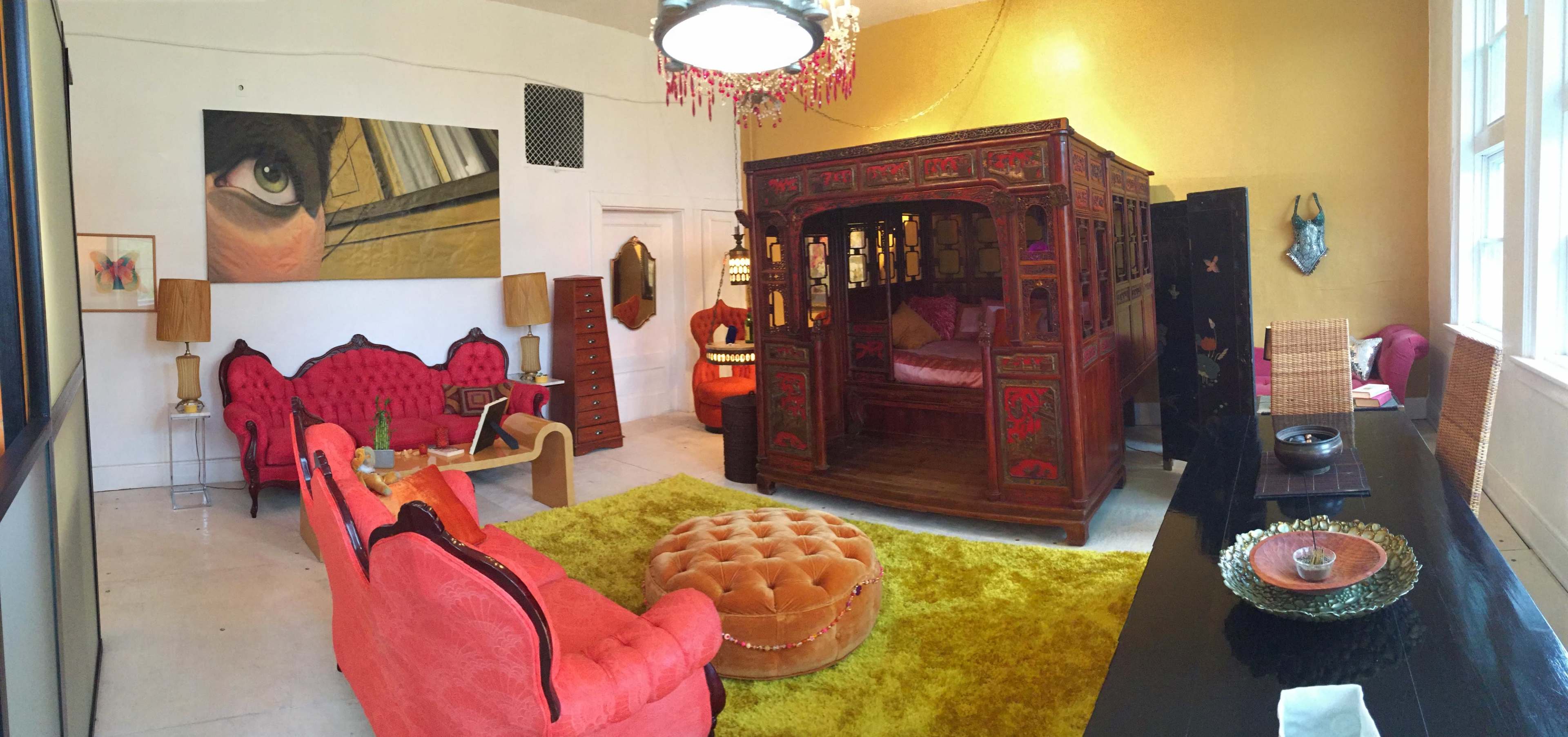 The scene shows a colorful living room featuring ornate red furniture, a vibrant yellow wall, a large wooden bedframe, and various decorative elements.