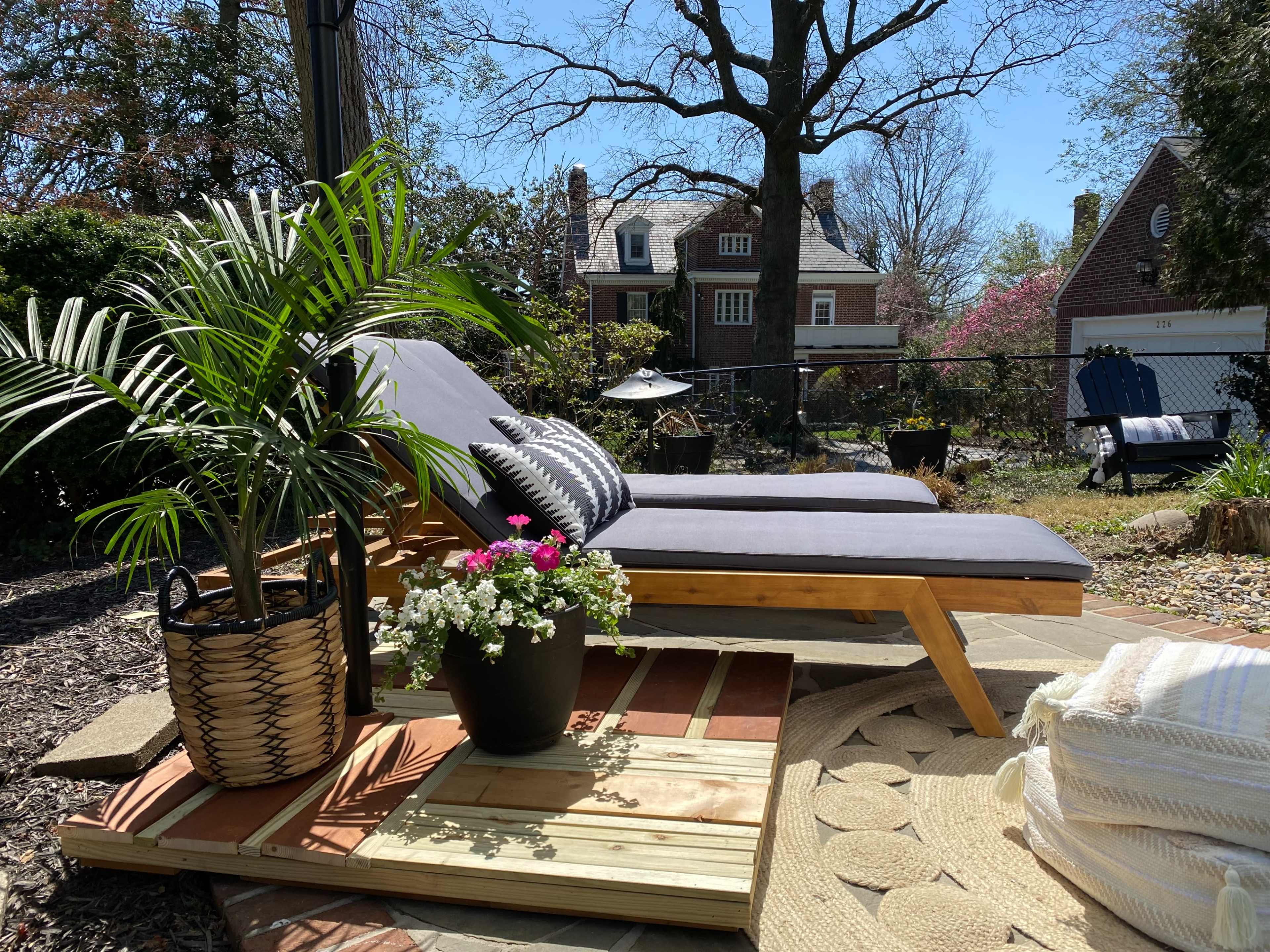 Two lounge chairs with gray cushions are positioned on a wooden patio surrounded by potted plants and a landscaped yard.