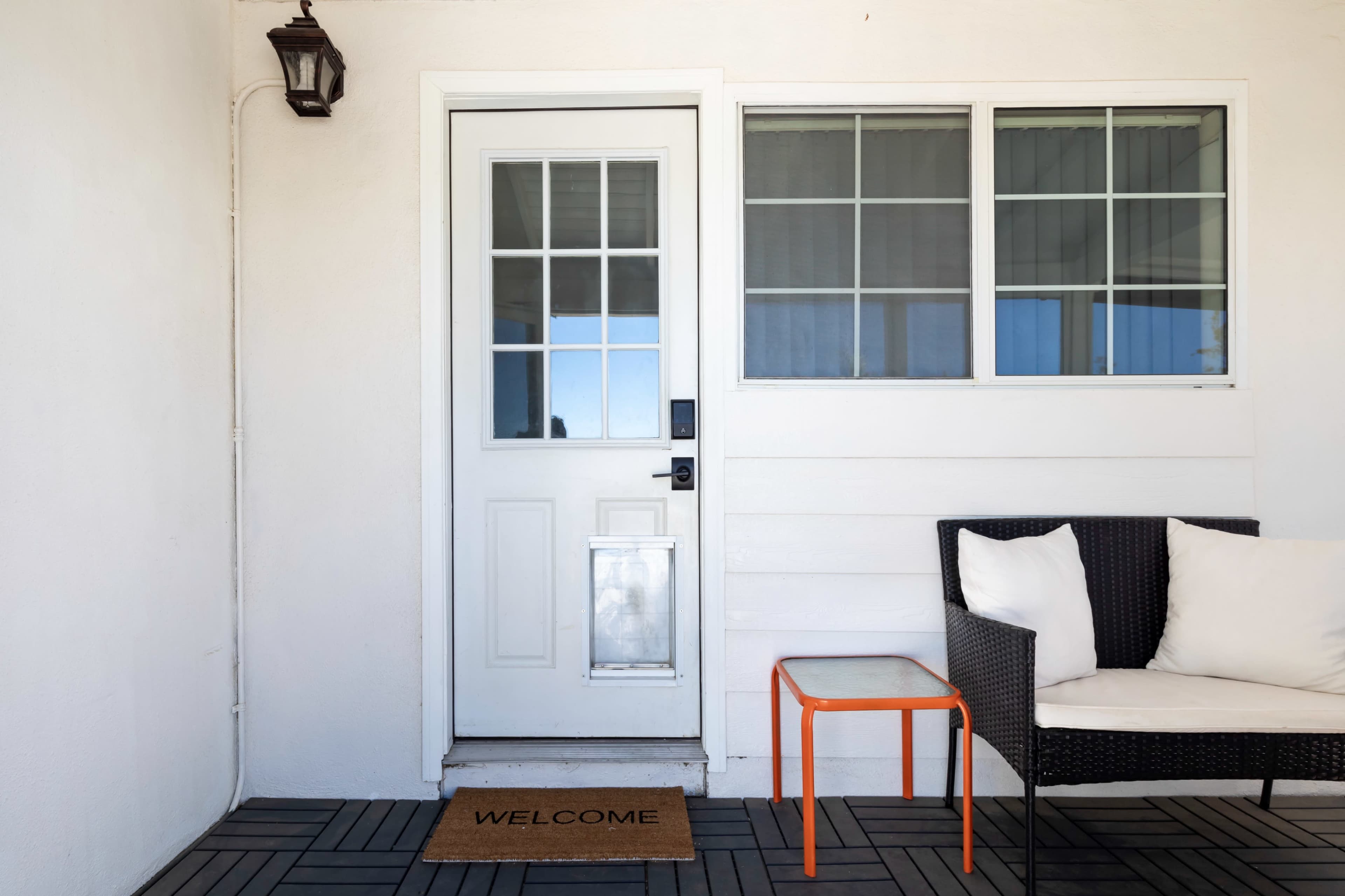 The image shows a front porch with a white door, a welcome mat, a small red table, and a black wicker chair.