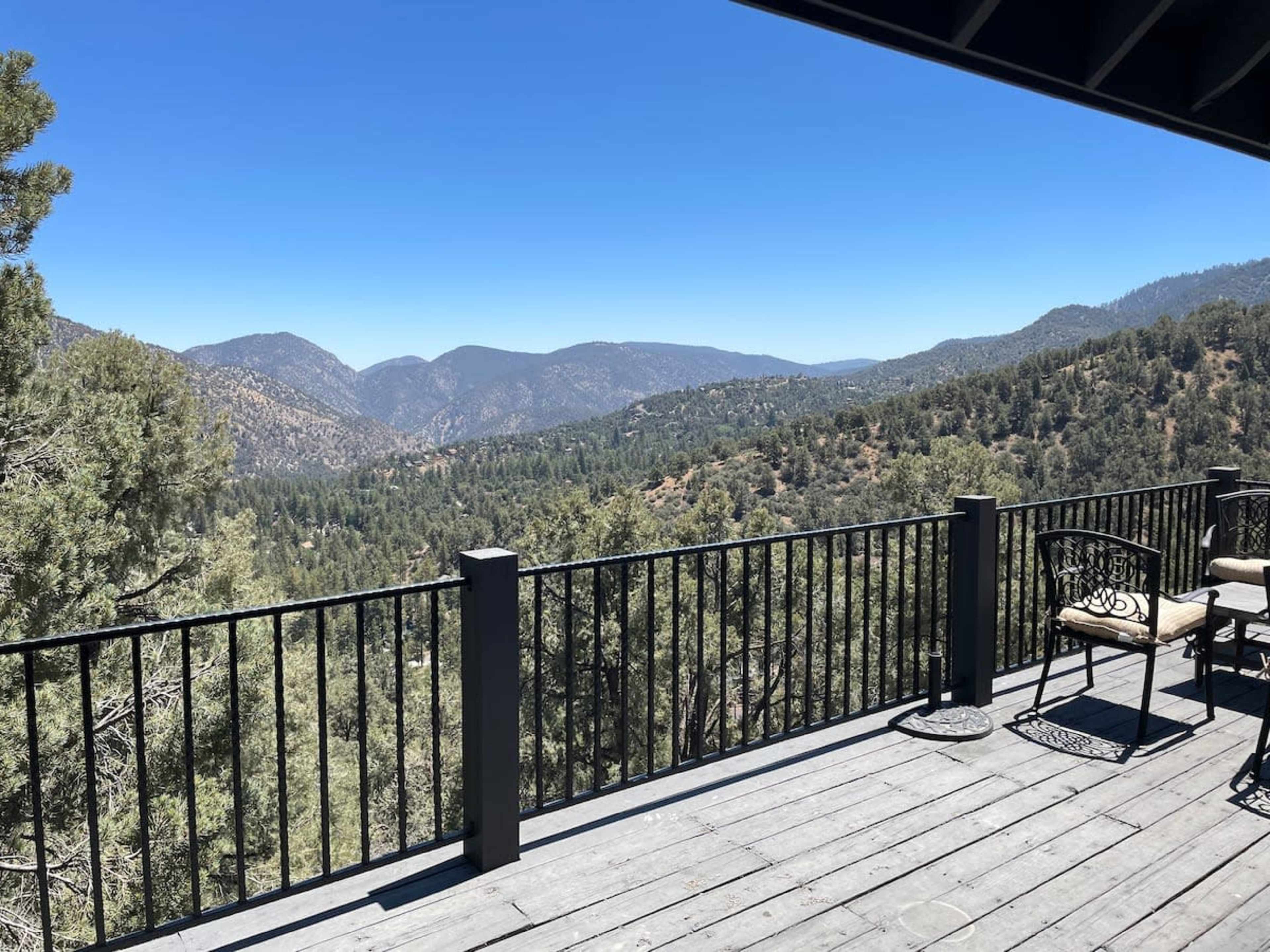A deck overlooks a mountainous landscape filled with trees and rolling hills under a clear blue sky.