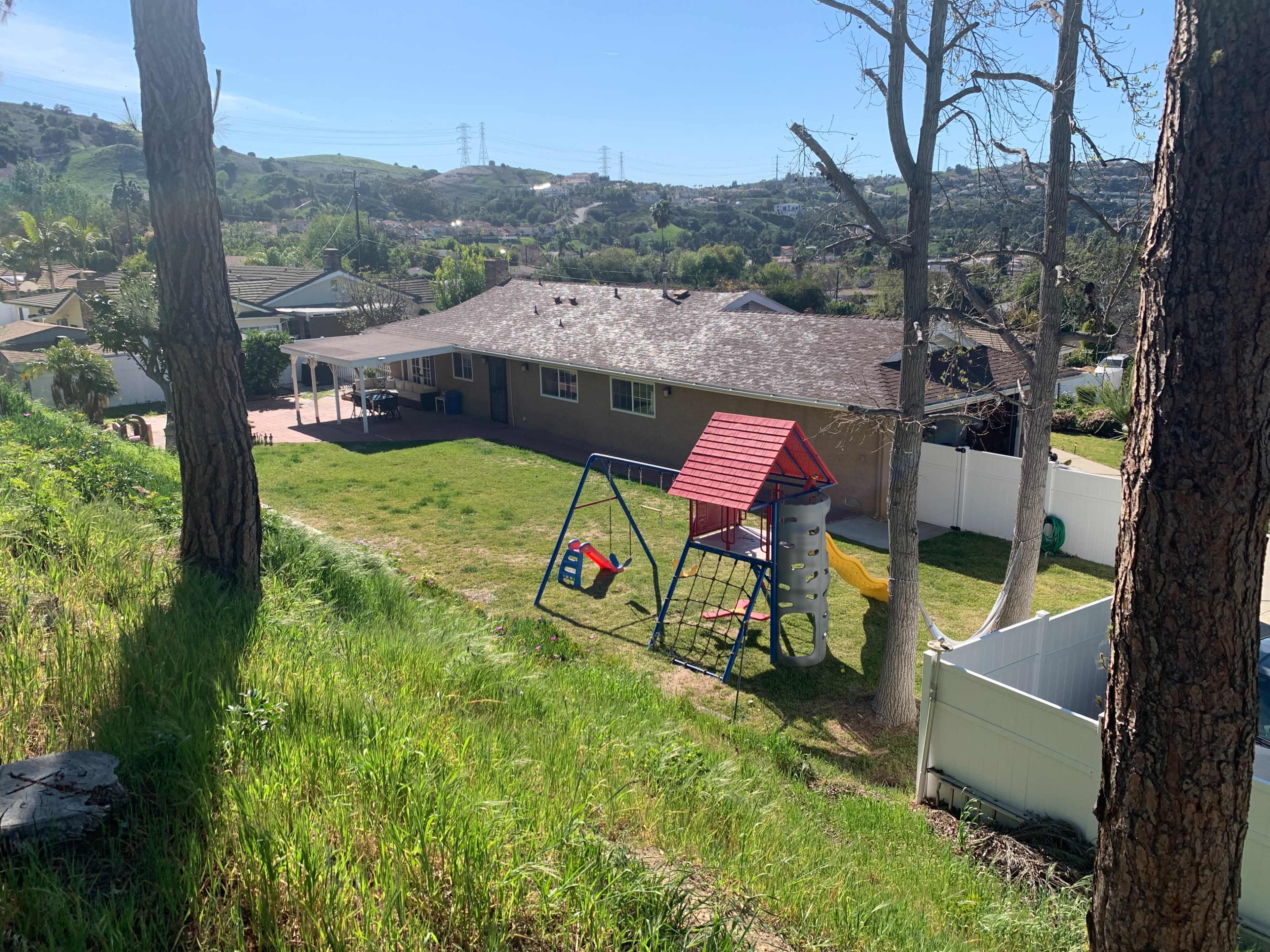 A backyard features a playset with a swing and slide, surrounded by grass and trees, with hills in the background.