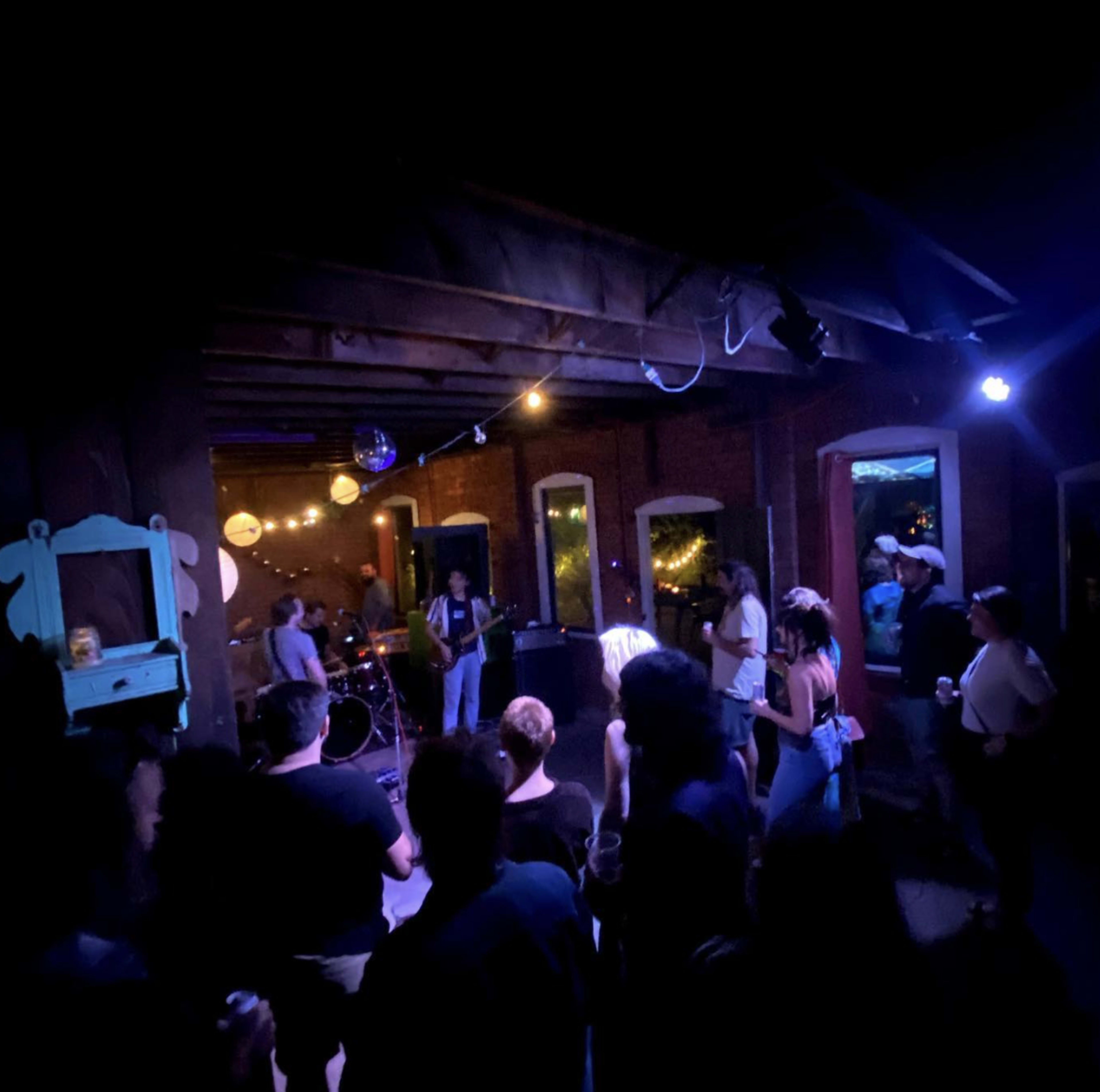 A small crowd gathers in a dimly lit venue with a live band performing on stage.