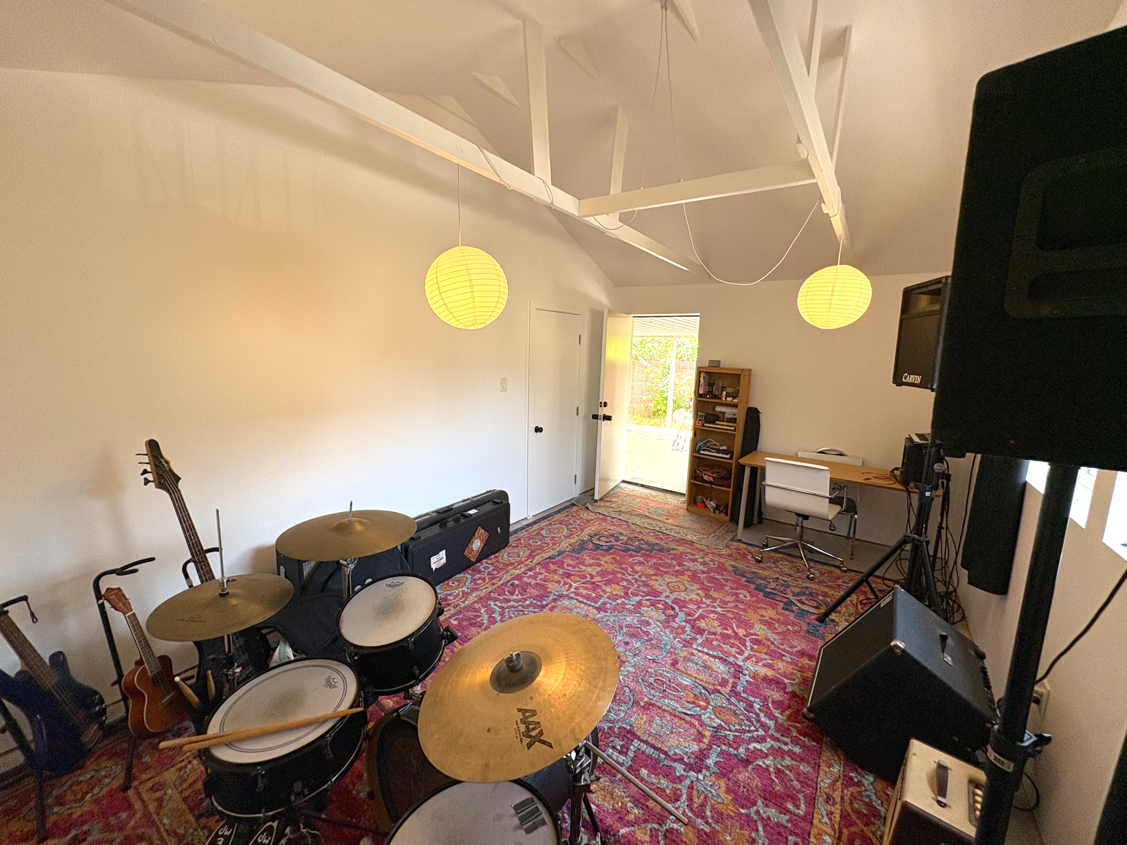 The image shows a music rehearsal space with a drum set, guitars, a computer desk, and a door leading outside.