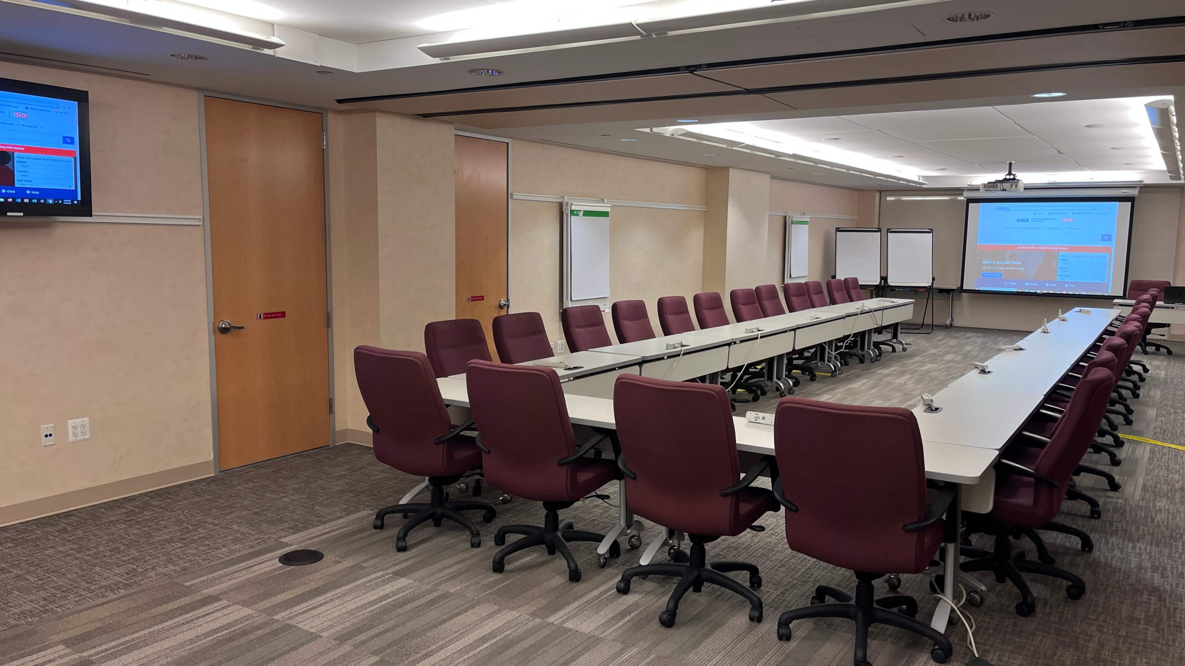 The image shows a conference room with a long table surrounded by maroon office chairs, screens displaying information, and whiteboards against the walls.