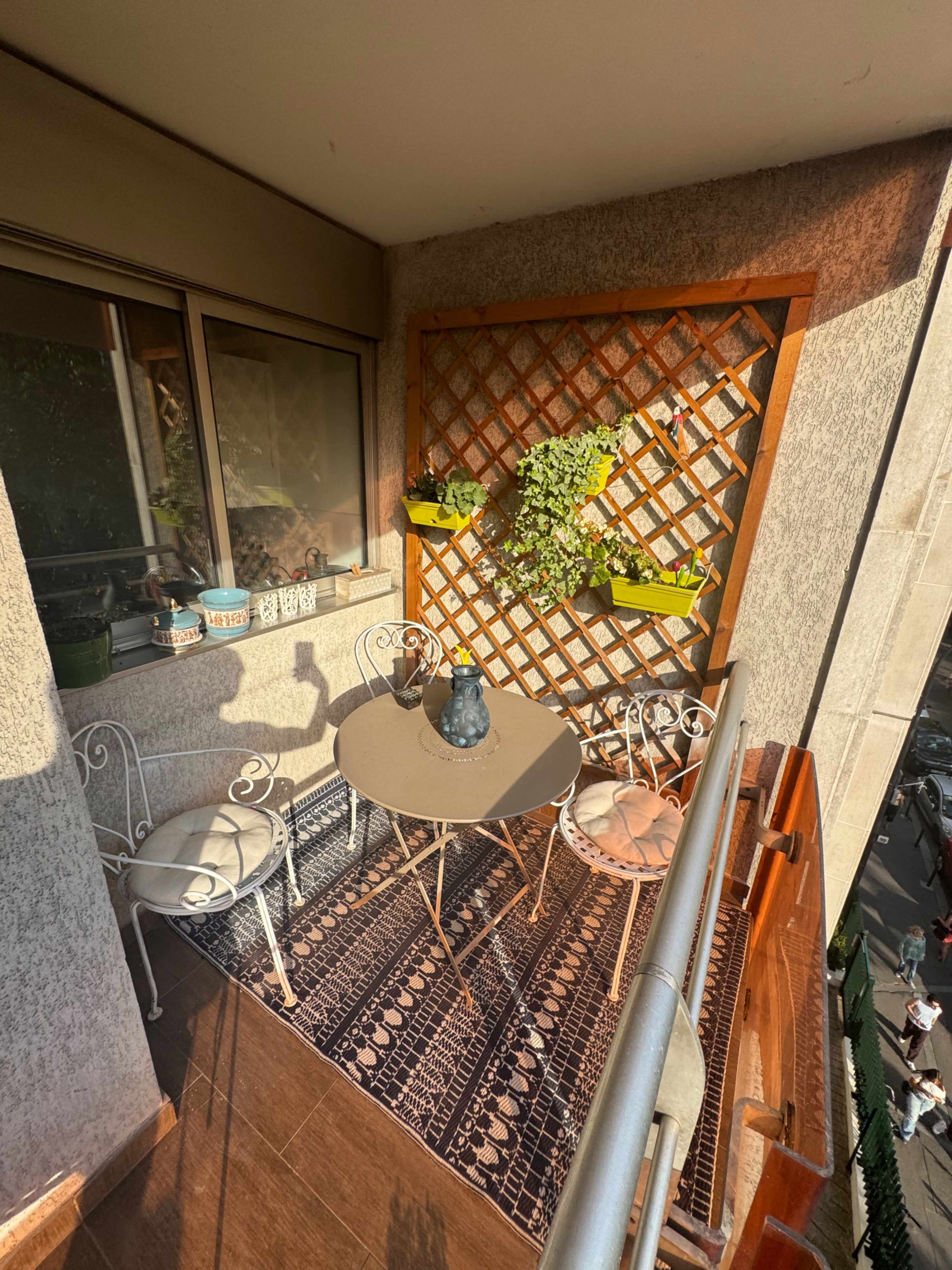 The image shows a balcony with a small table, four chairs, and a wooden lattice adorned with plants.