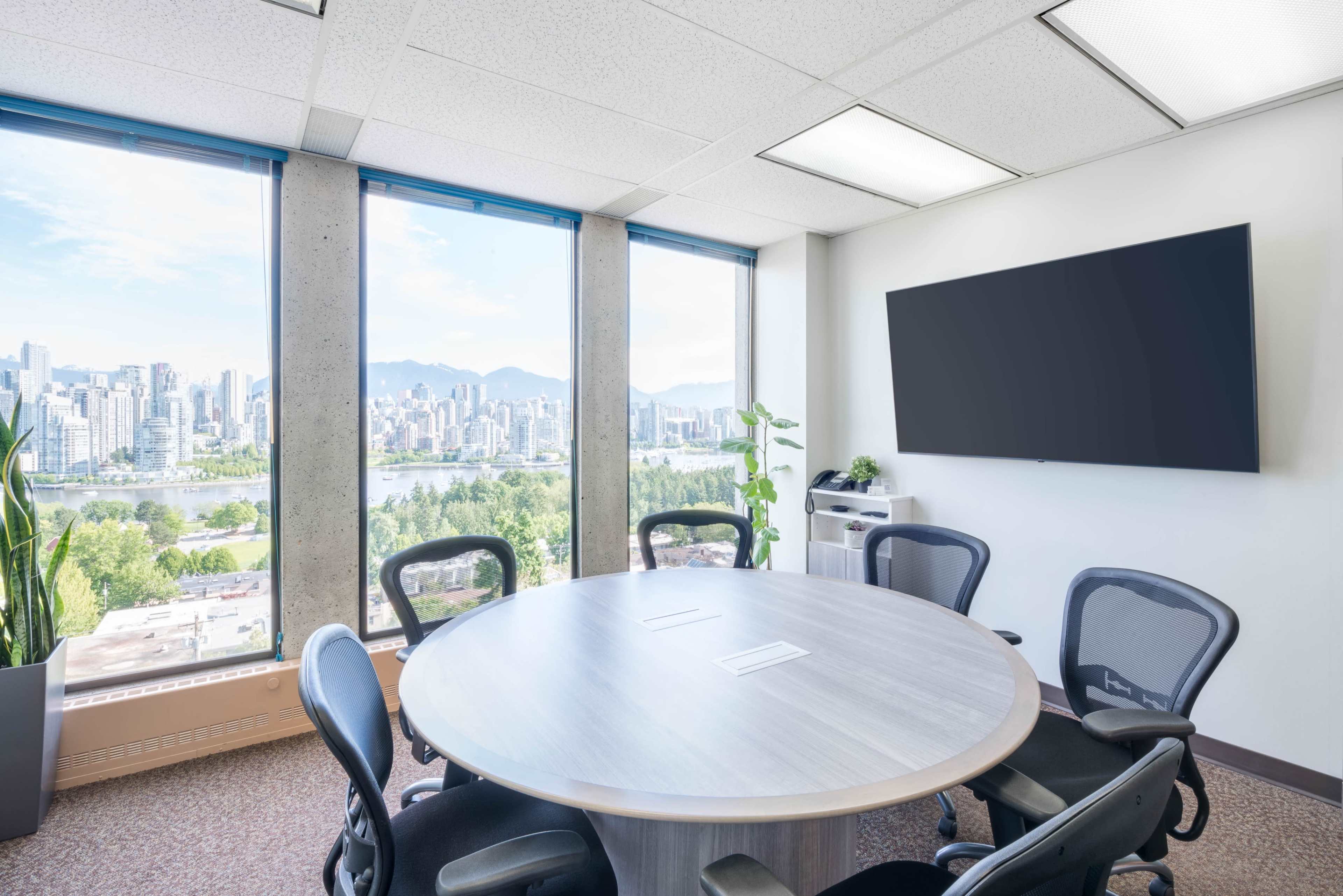 A modern conference room features a round table surrounded by office chairs, with large windows offering a view of a city skyline and mountains.