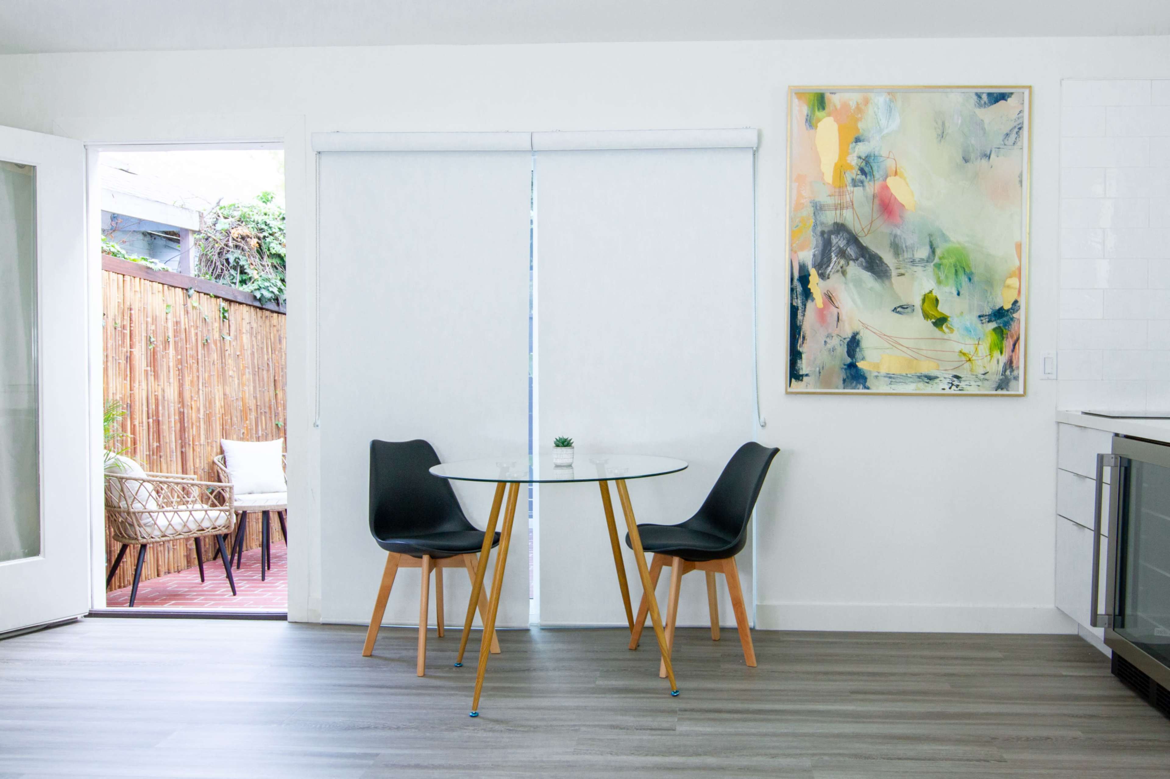 A small dining area features a round glass table with two black chairs and a large colorful painting on the wall, next to an open sliding door leading to a private outdoor space.