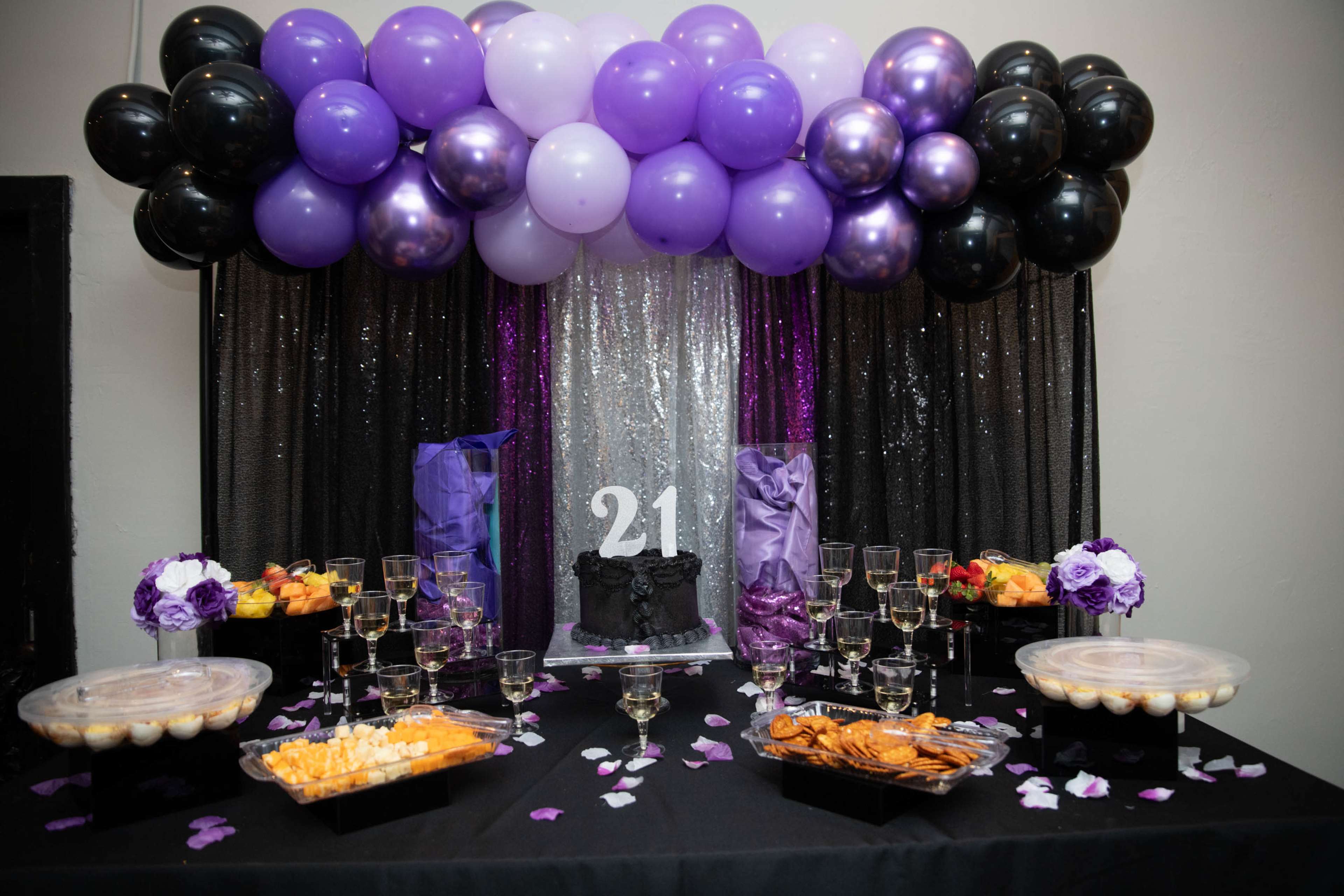 A table is decorated with a black and purple theme, featuring a cake with "21," surrounded by cupcakes, cookies, and elegant glassware, under a backdrop of balloons and sparkling drapes.