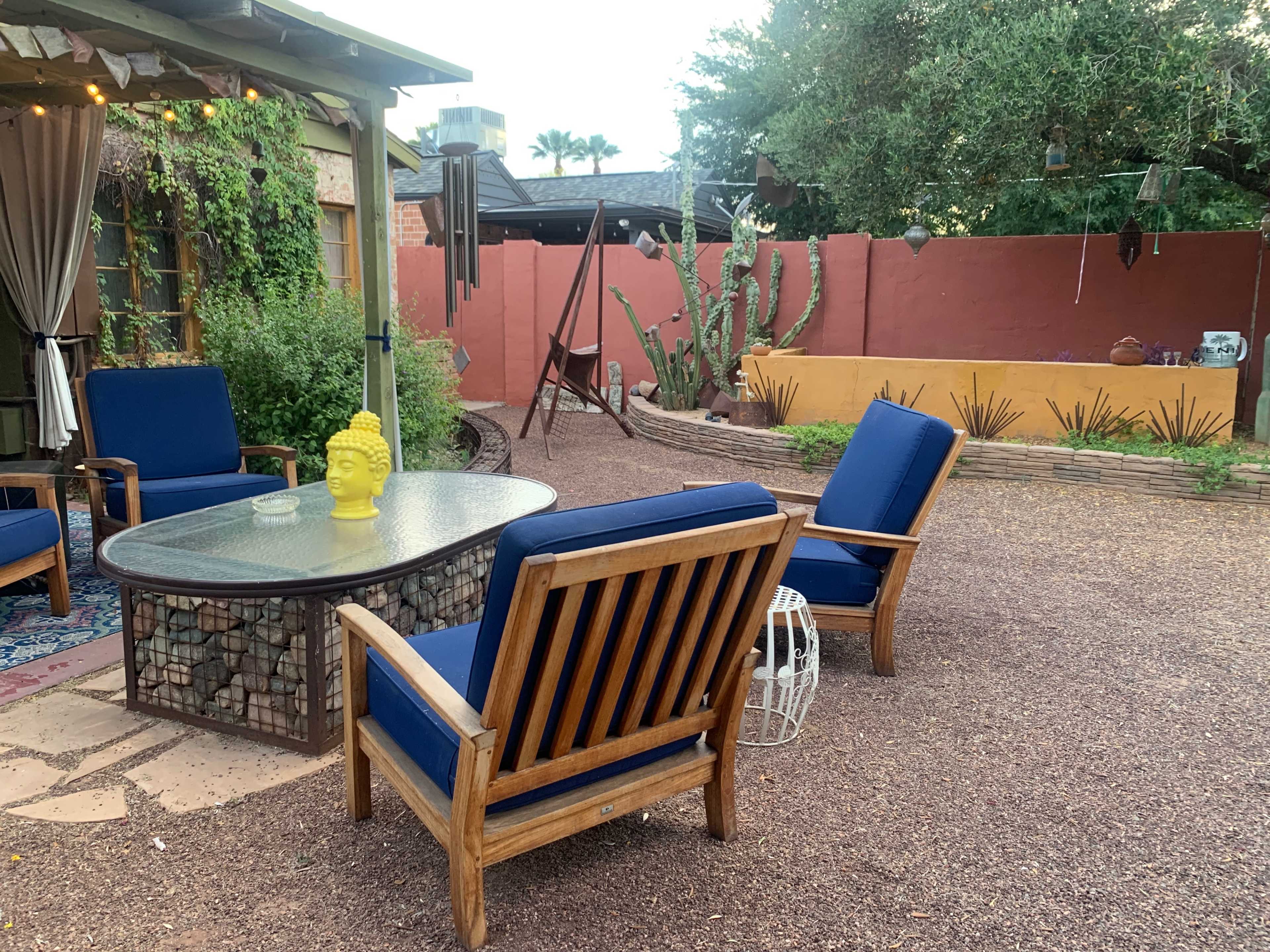 The scene features a backyard patio with blue cushioned wooden chairs surrounding a glass-top table, set against a backdrop of desert plants and a colorful wall.