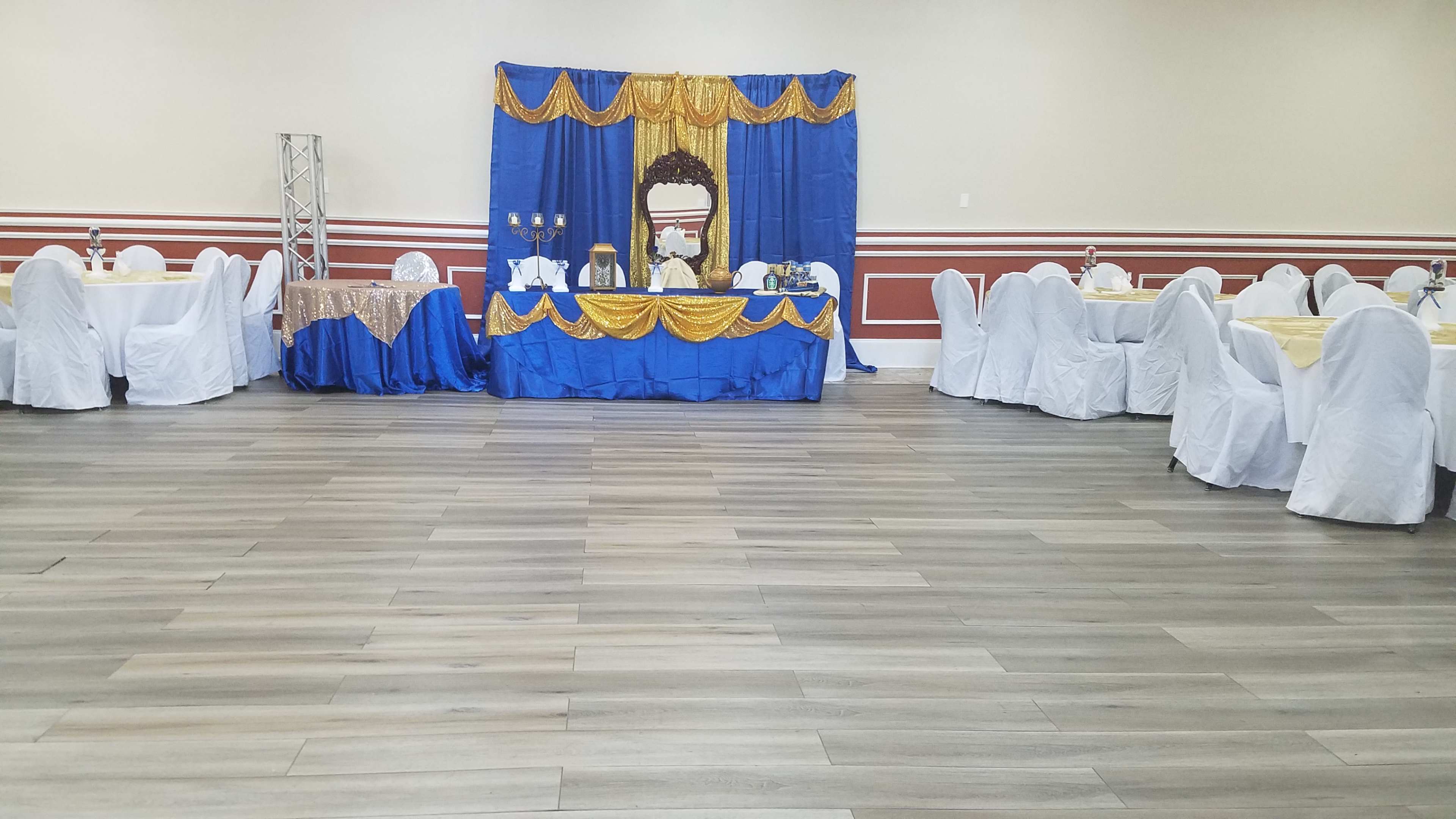 The image shows a decorated banquet hall with white tablecloths and a backdrop of blue and gold fabric featuring a mirror and various decorative items on a table.