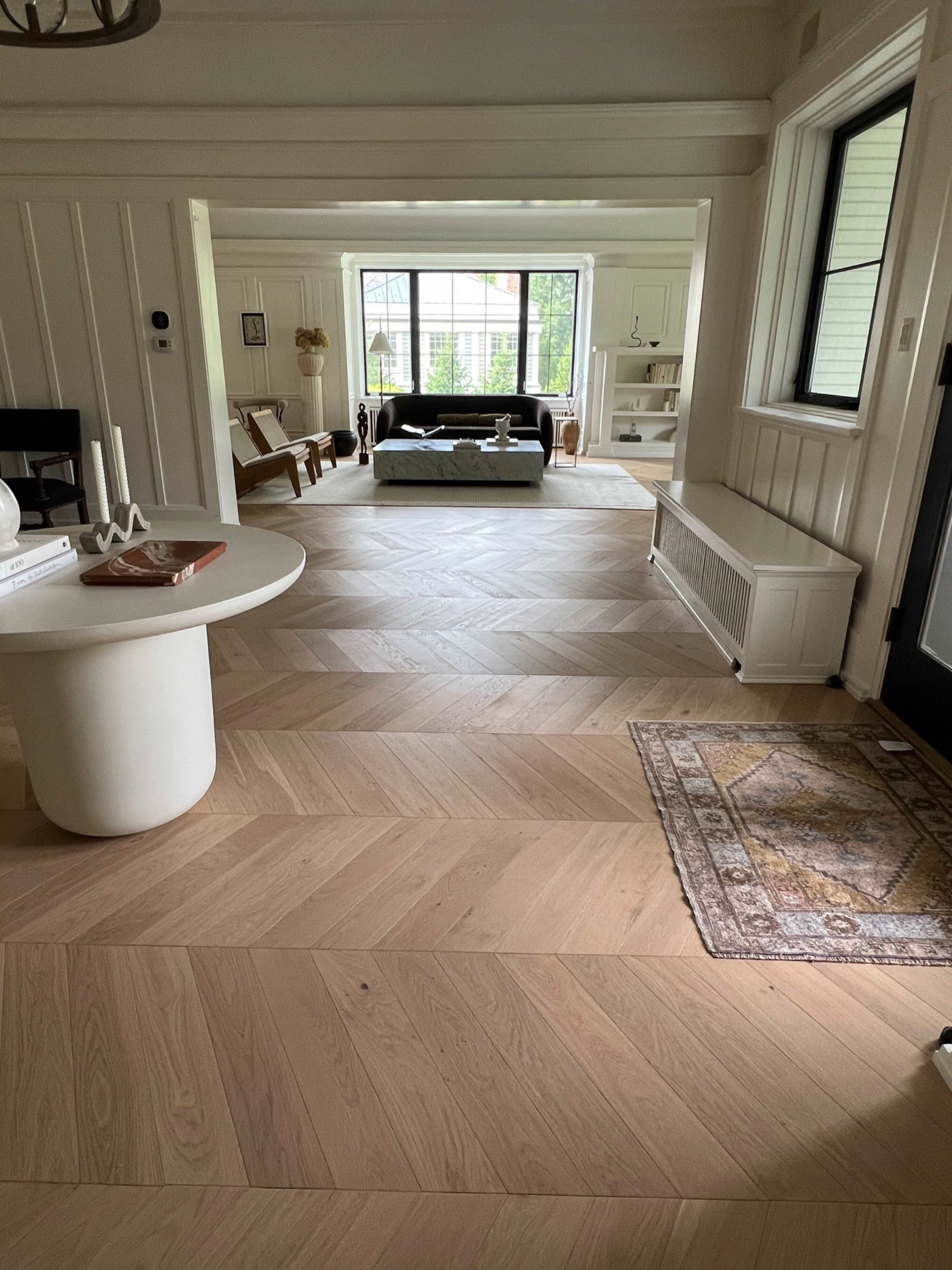 A spacious interior with a herringbone-patterned wooden floor, leading to a living area that features a couch and a large window.