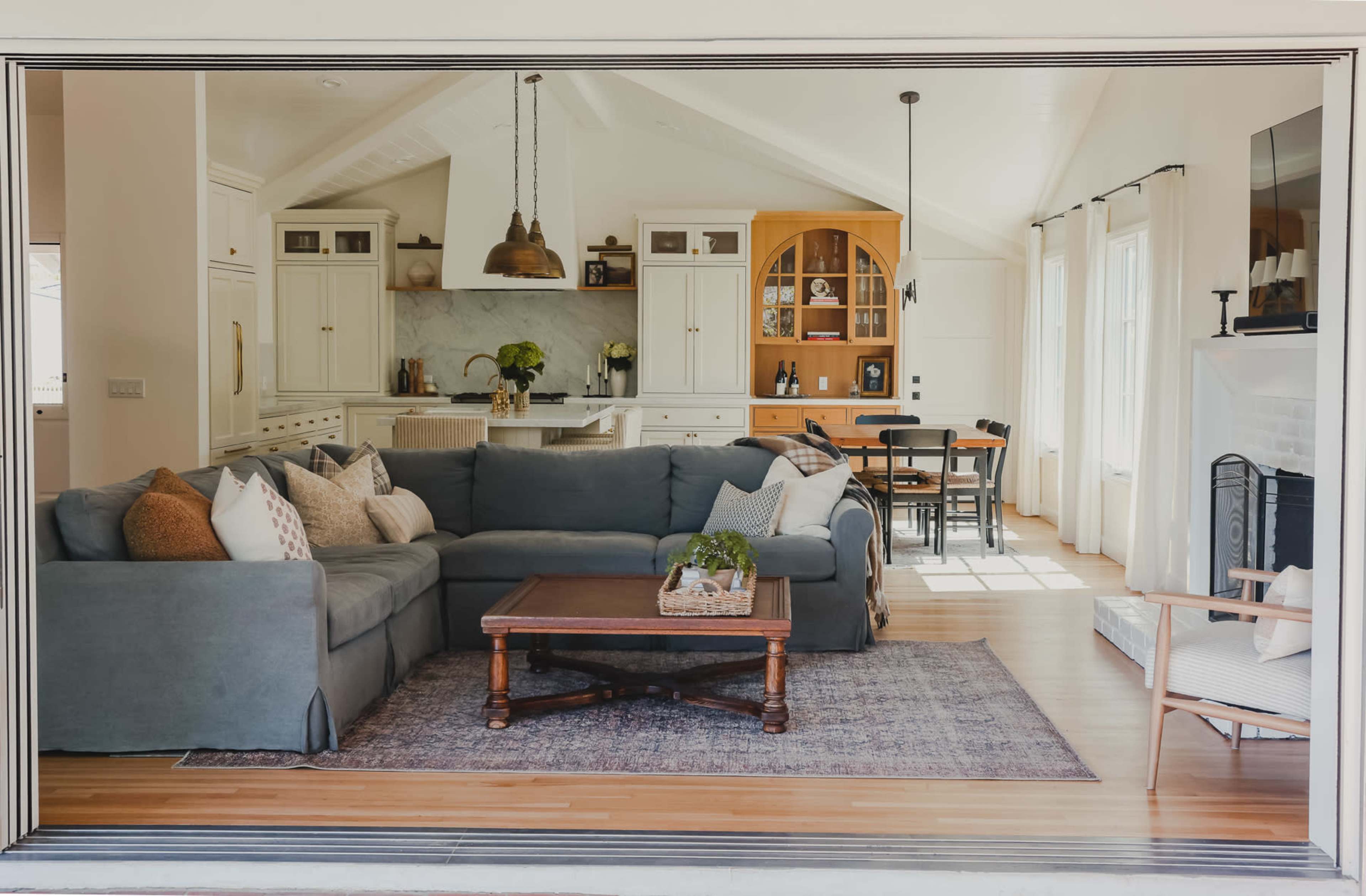 A bright, open living space featuring a gray sectional sofa, a wooden coffee table, and a modern kitchen in the background.