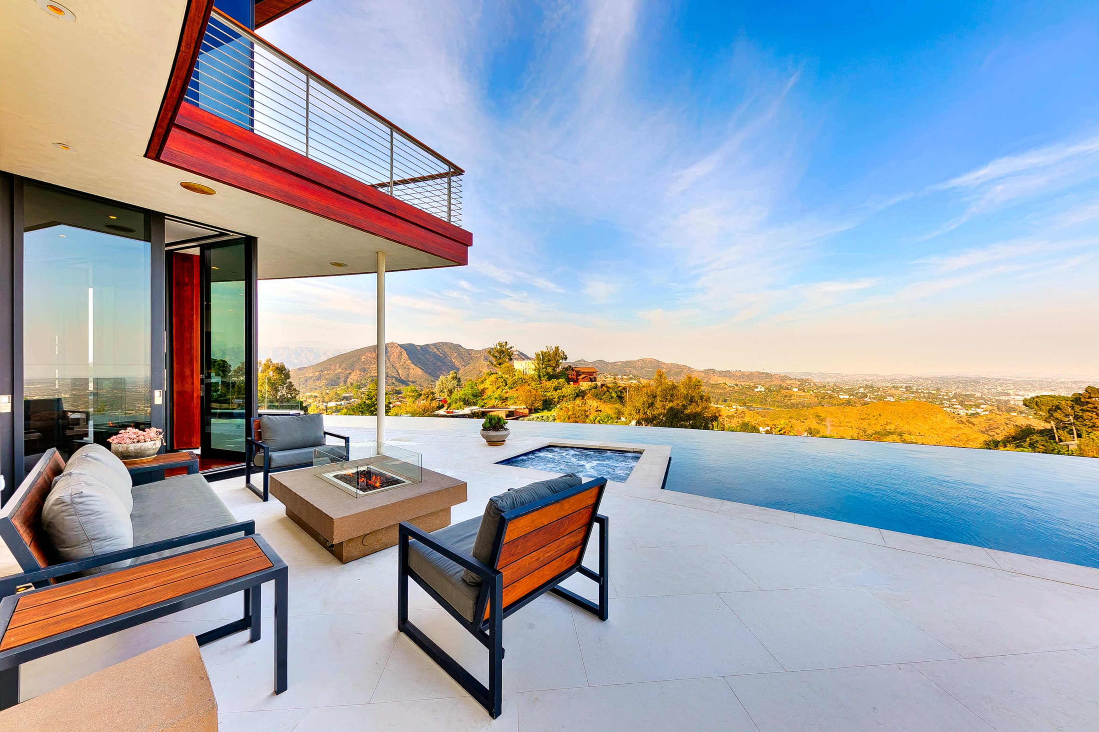 The image shows a patio with modern furniture overlooking a pool and mountainous landscape under a clear blue sky.