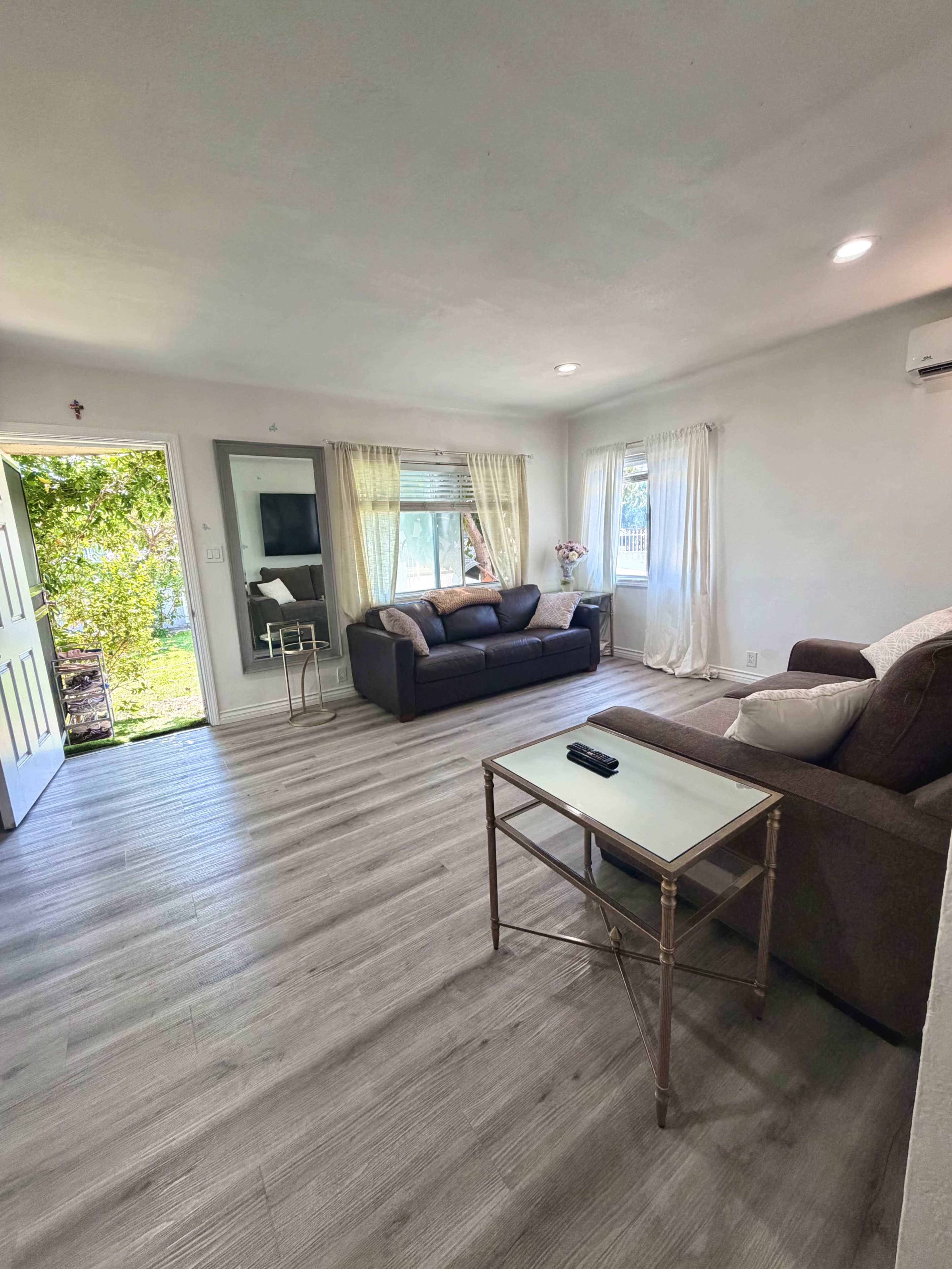 The image shows a living room featuring two sofas, a coffee table, and large windows that provide natural light and an outdoor view.