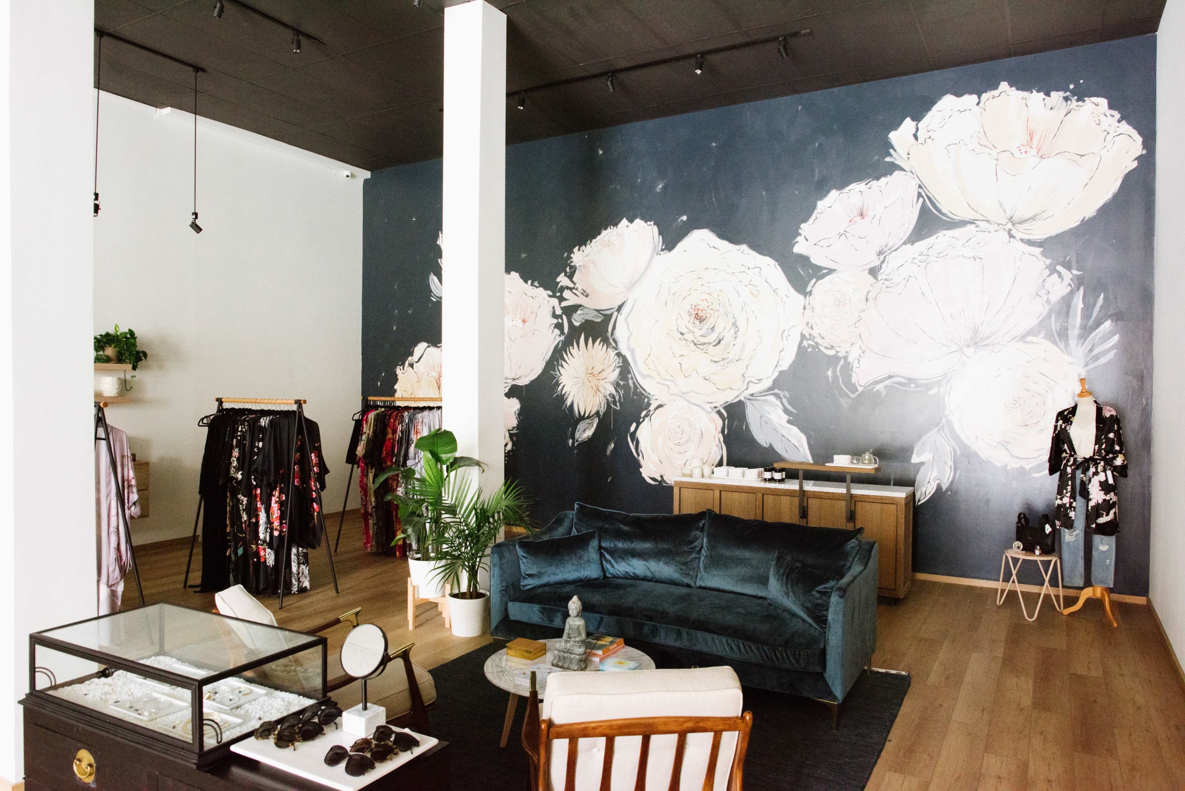 The scene shows a boutique with a large floral mural on the wall, clothing racks on the left, a styled seating area with a velvet couch in the center, and a display case in front.