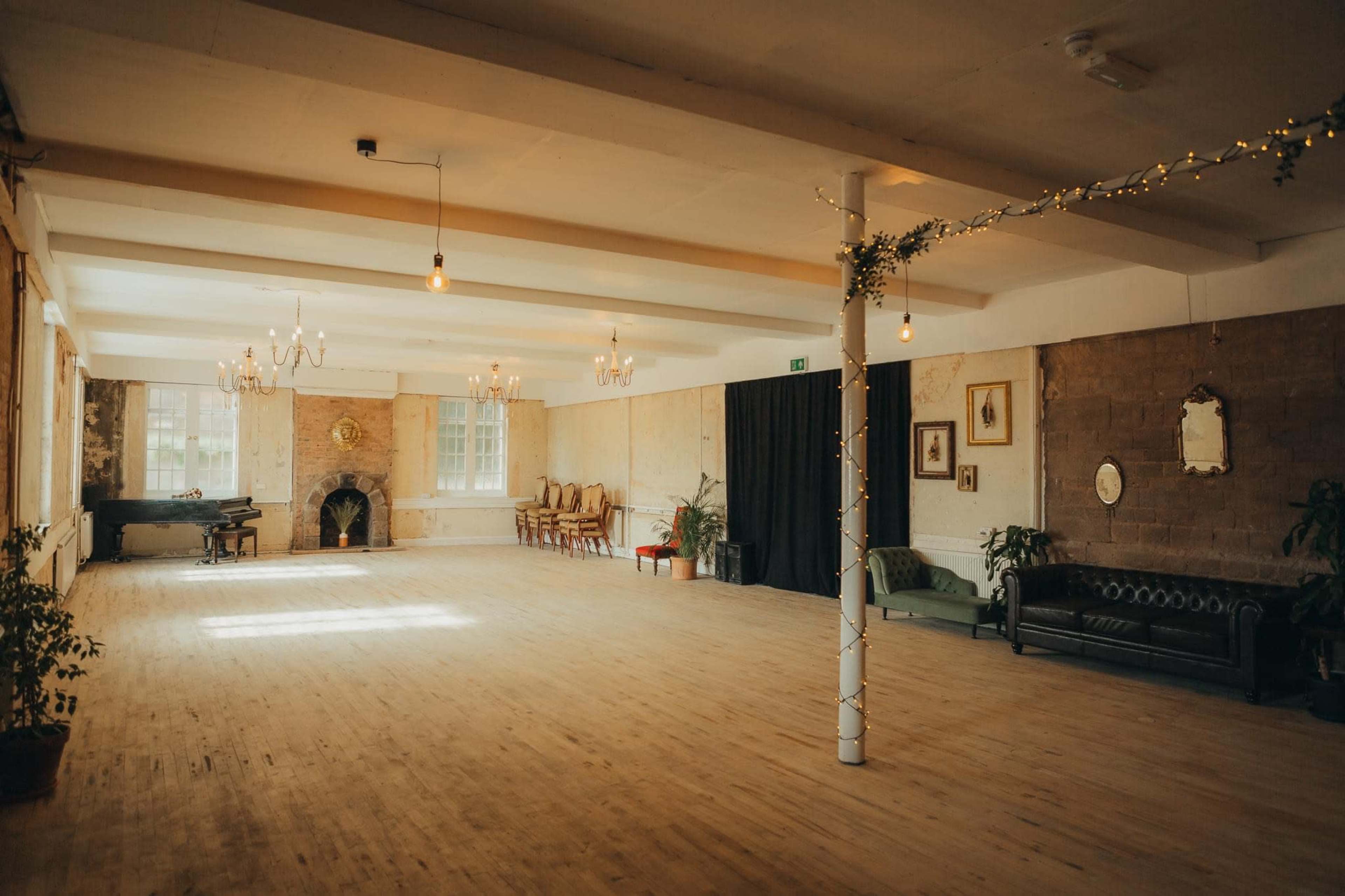 A spacious, well-lit room with wooden flooring, decorated with plants, a fireplace, and vintage furniture.