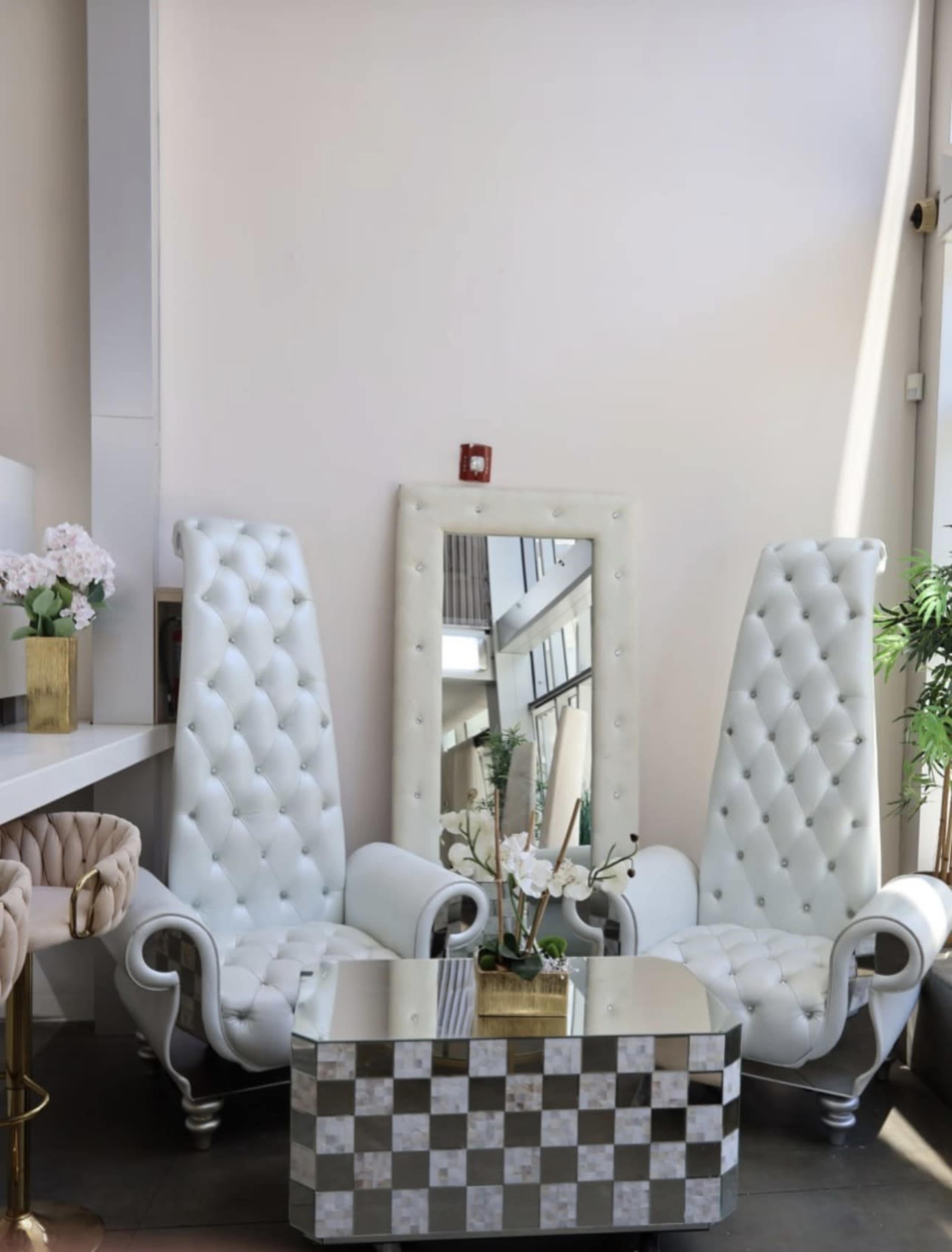 The image shows a stylish seating area featuring two tall, white tufted chairs flanking a mirrored table with a checkered design, set against a light-colored wall.