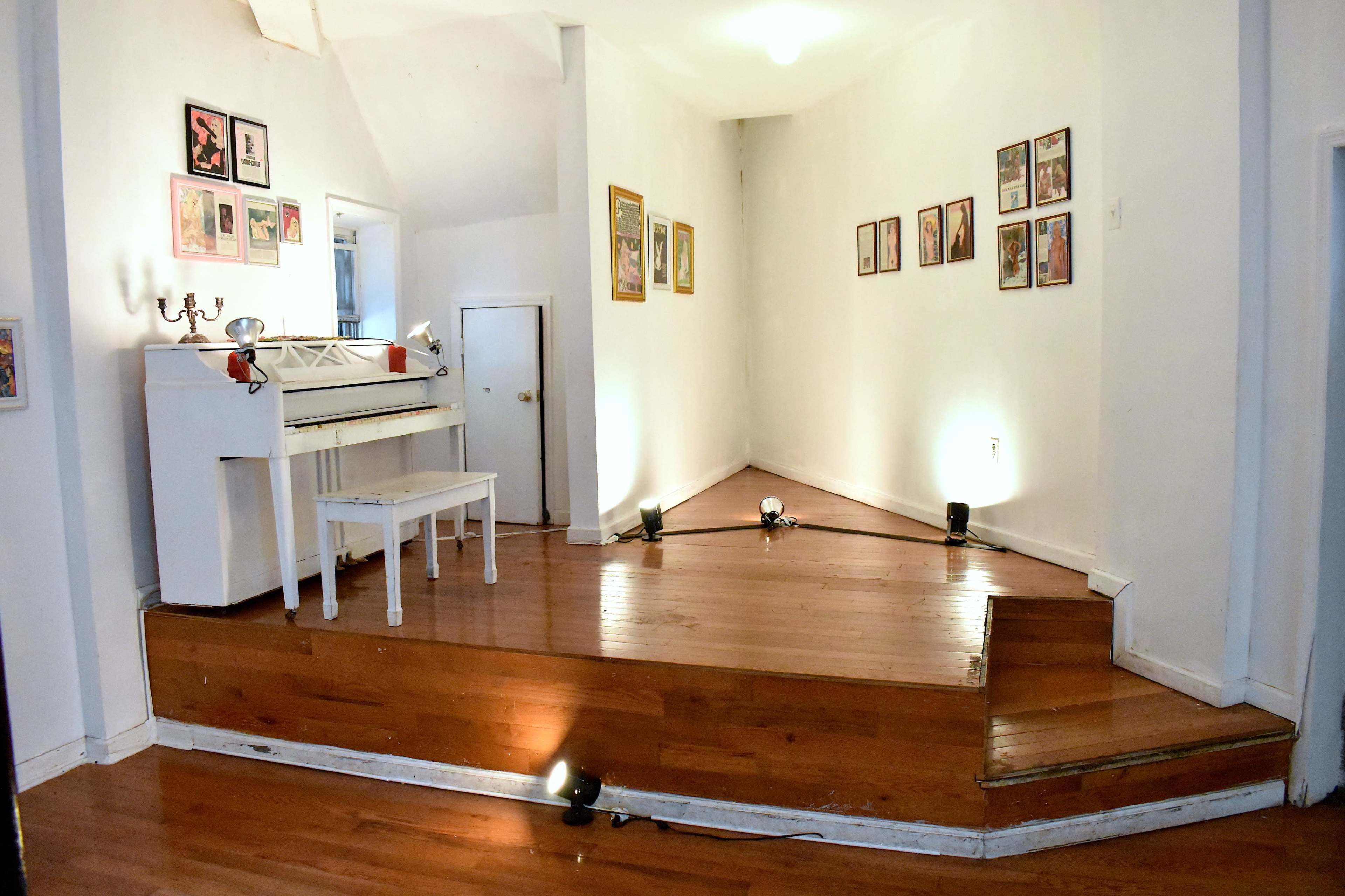 A bright, minimalist room featuring a white piano and a small stool on a wooden platform, with framed pictures arranged on the walls.