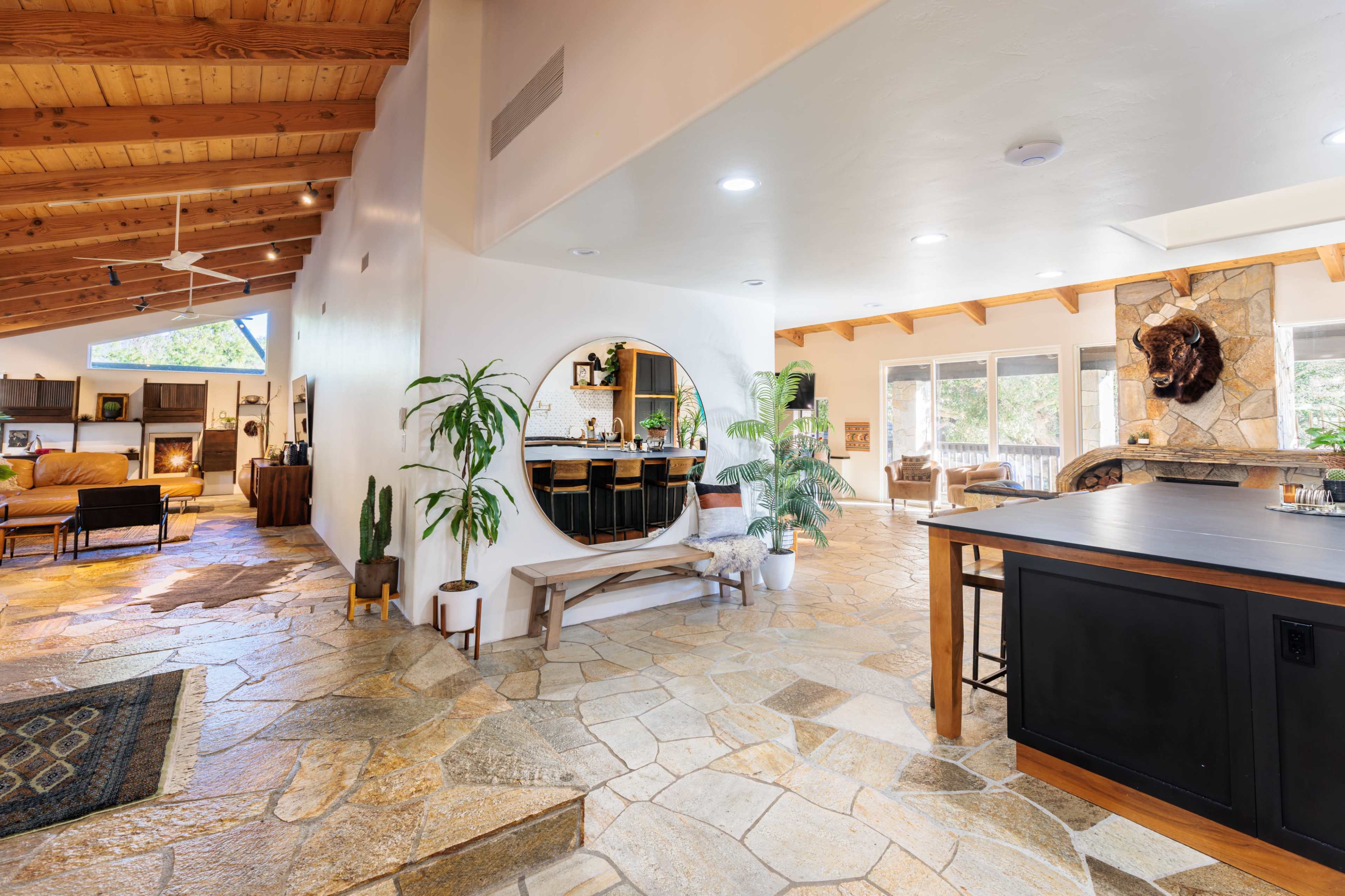 The image shows an open-concept living space featuring natural stone flooring, a circular mirror, a spacious kitchen island, and large windows allowing ample light.