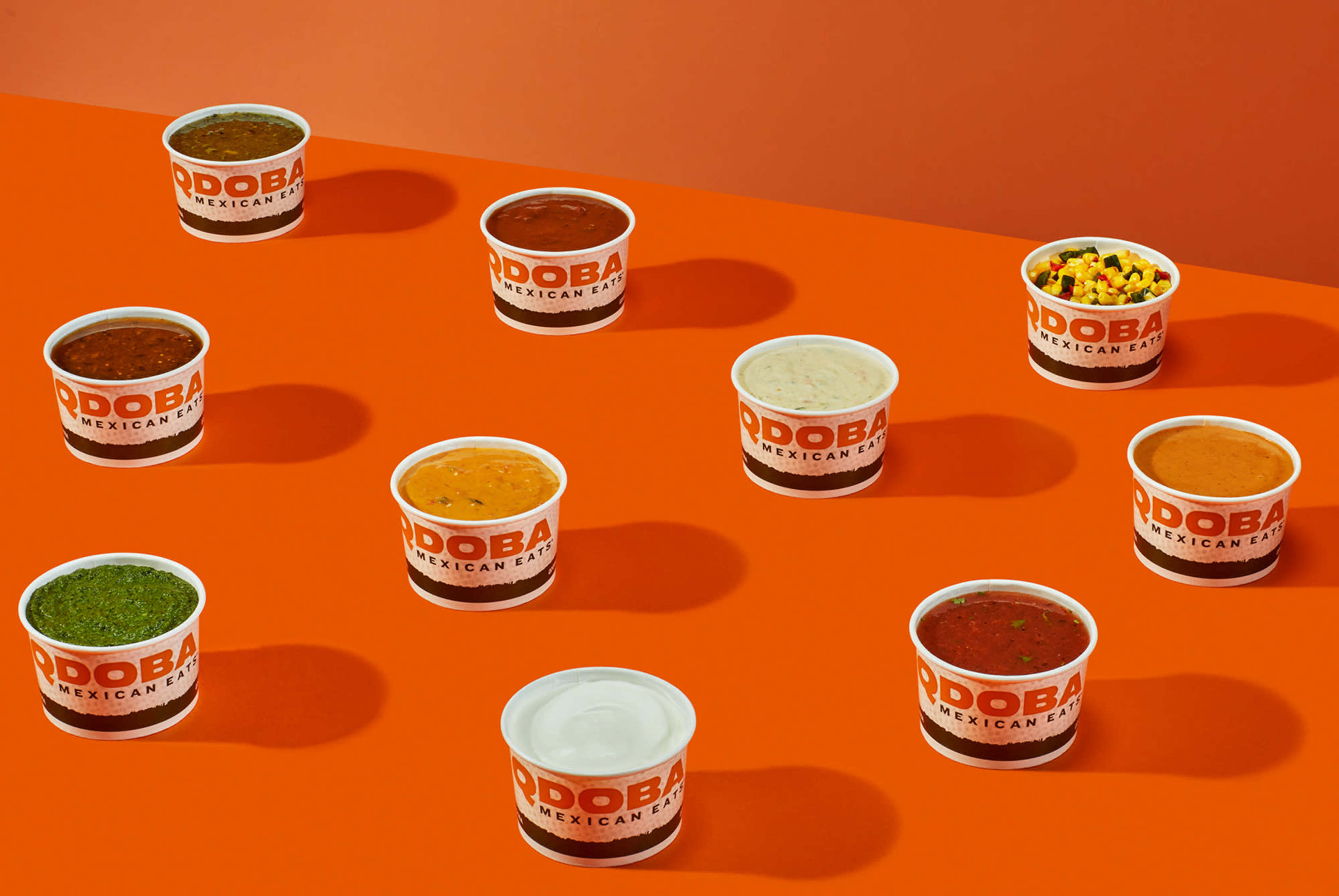 The image features an arrangement of ten containers of various sauces and toppings from QDOBA on a bright orange background.