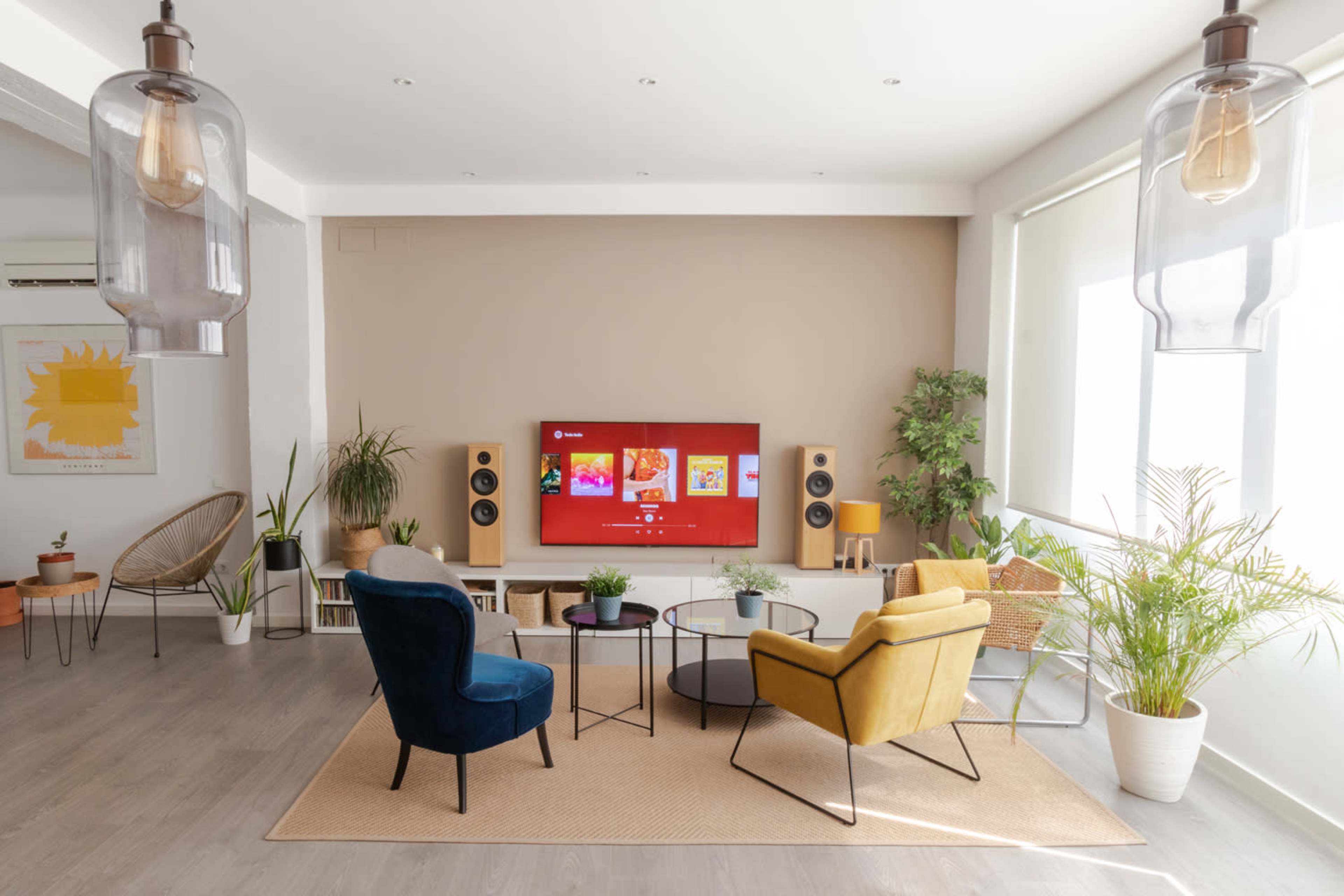 A modern living room features two chairs, a coffee table, and a large TV mounted on the wall, surrounded by plants and decorative items.