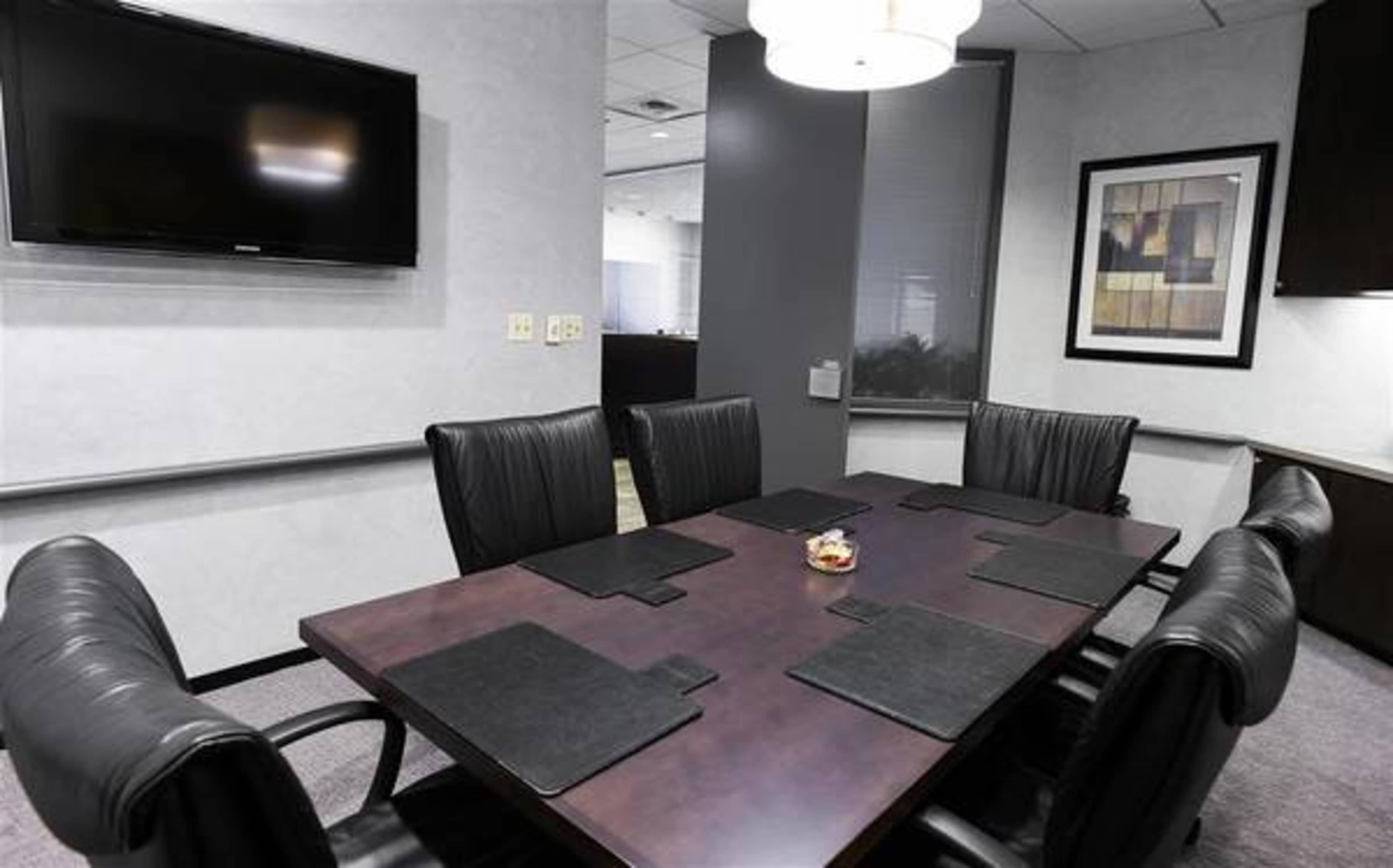 A conference room with a large table surrounded by leather chairs, a television mounted on the wall, and a decorative piece framed on the adjacent wall.