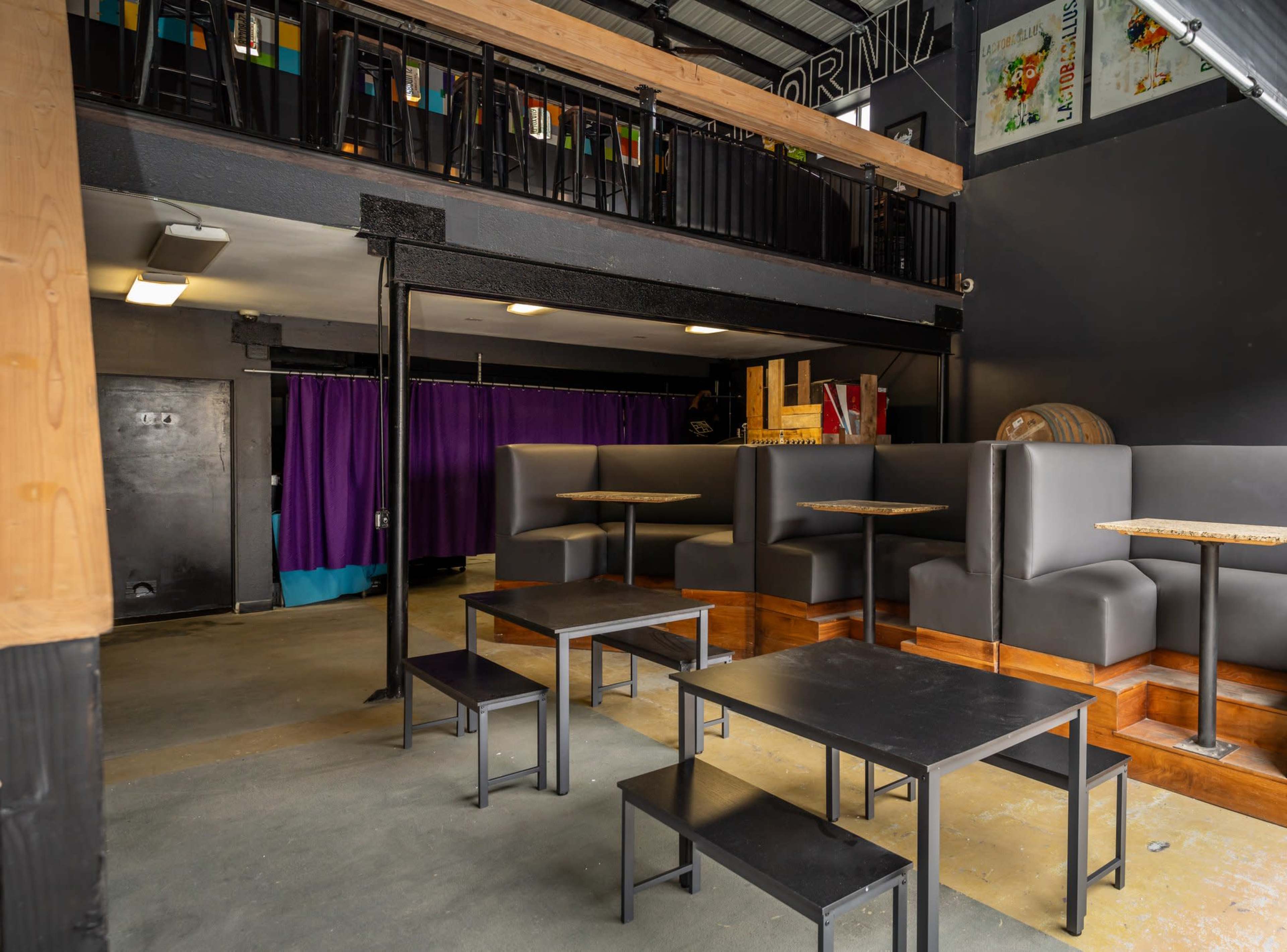 A modern café interior with dark tables and booths, a purple curtain in the background, and a mezzanine with colorful chairs.