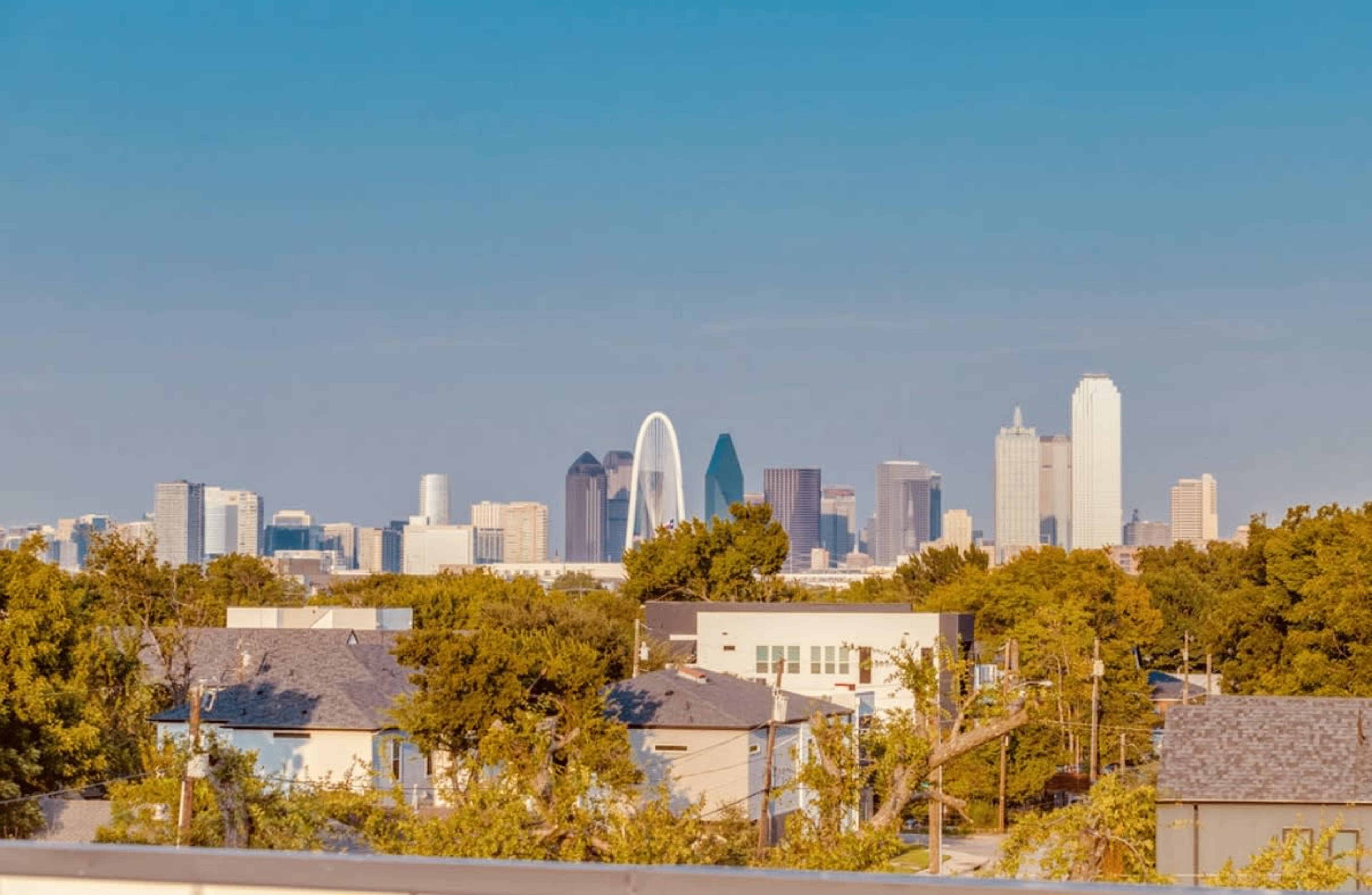 The image shows a skyline view of Dallas, Texas, featuring a mix of modern skyscrapers and a large arch structure, with trees and rooftops in the foreground.